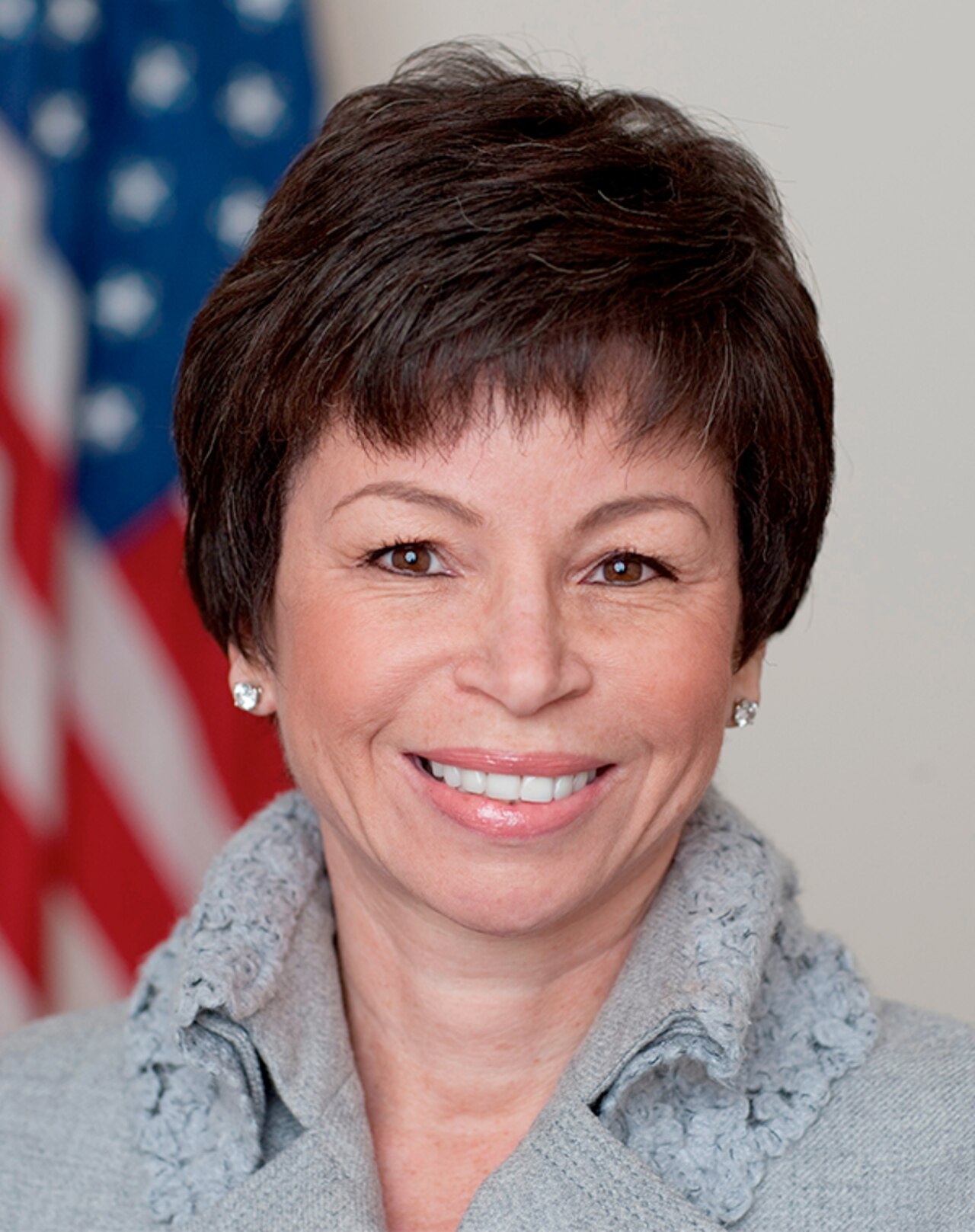 Valerie Jarrett smiles, wearing a grey sweater and diamond earrings, with an American flag blurred in the background.