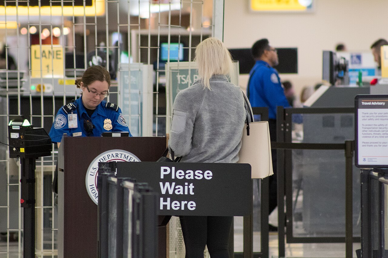 TSA officer in blue uniform at a podium with a sign that says Please Wait Here