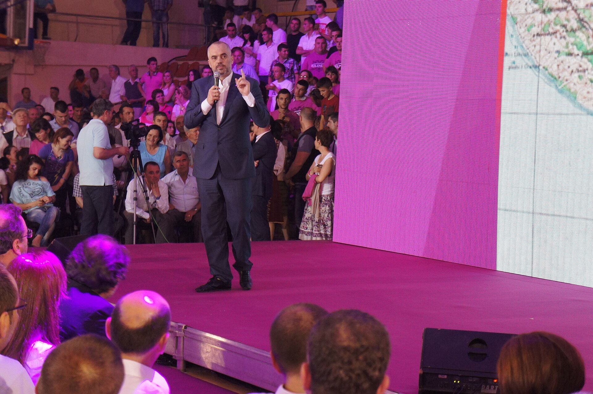 A man in a dark suit and white shirt, holding a microphone, stands on a pink stage and addresses a crowd. He is gesturing with his left hand. Behind him, a large pink screen displays a pattern of horizontal lines, and to the right, a screen shows a map.