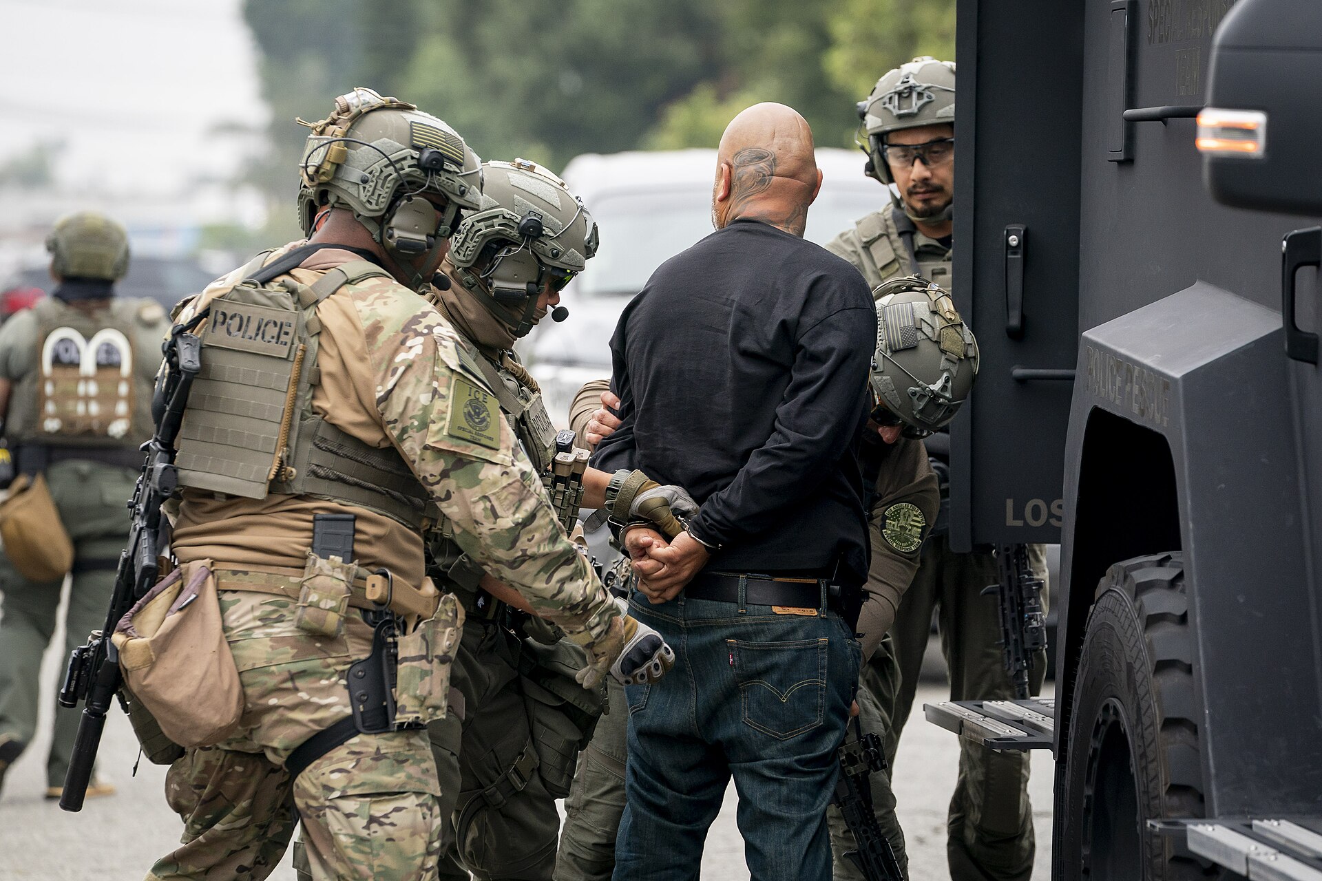 Several law enforcement officers in tactical gear are arresting a bald man with tattoos on his head. The officers have their hands on the man, who is wearing a black long-sleeved shirt and blue jeans, and appear to be handcuffing him. A large black vehicle, possibly an armored truck, is visible to t
