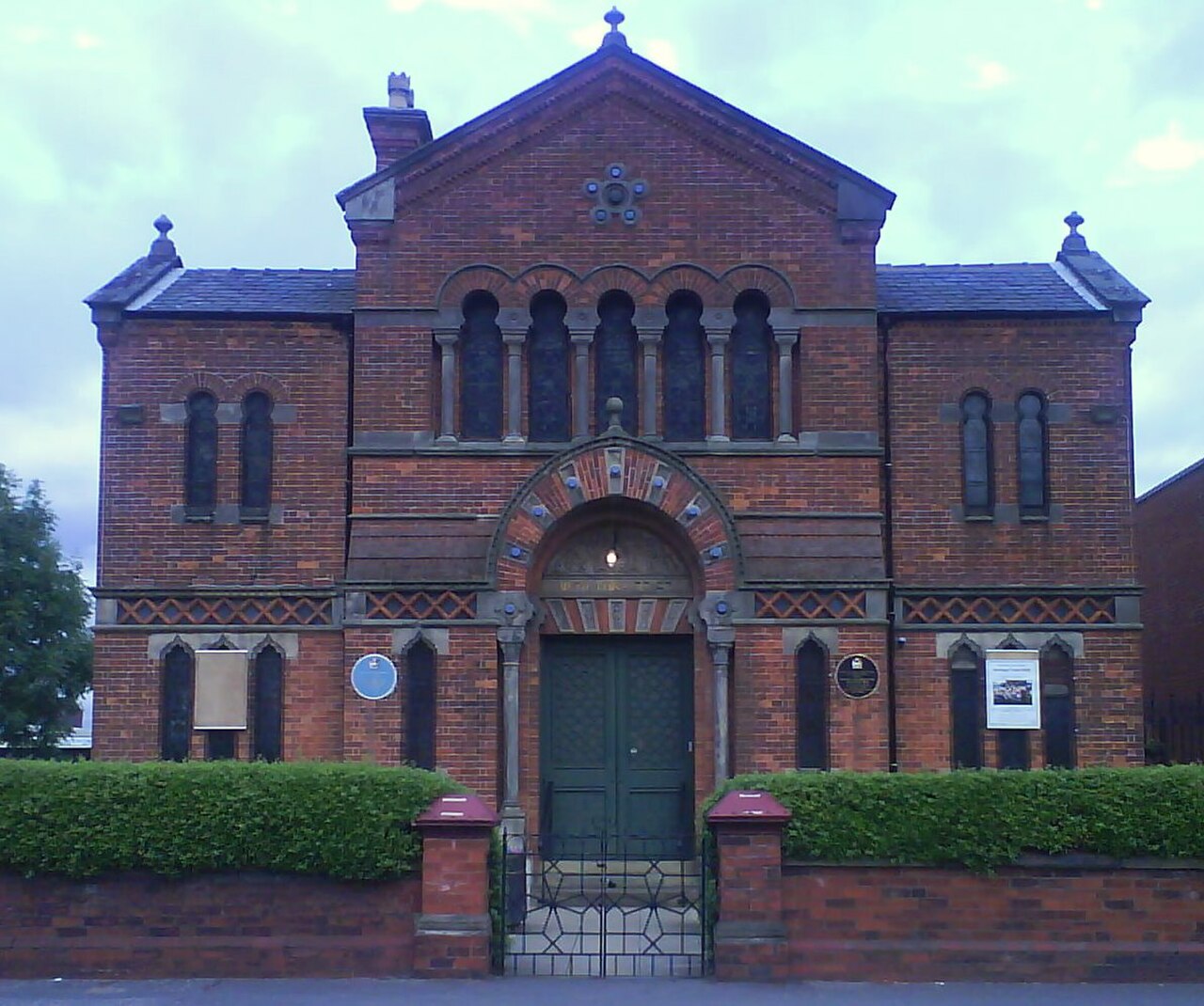 The Heaton Park synagogue, a brick building with a green door and arched entrance, surrounded by hedges and a gate.