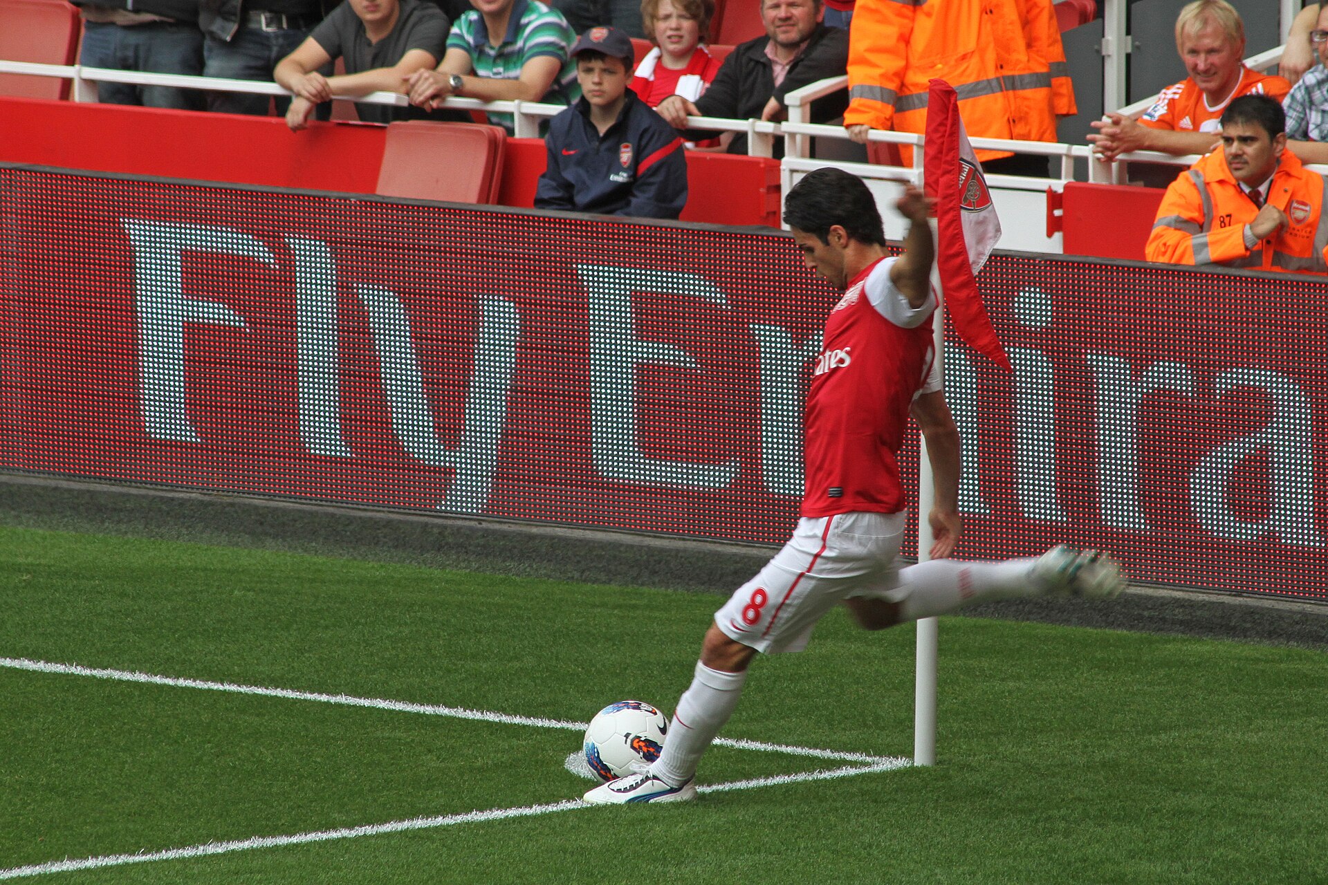 A soccer player in a red and white Arsenal jersey with the number 8 on his shorts kicks the ball from the corner of the field.