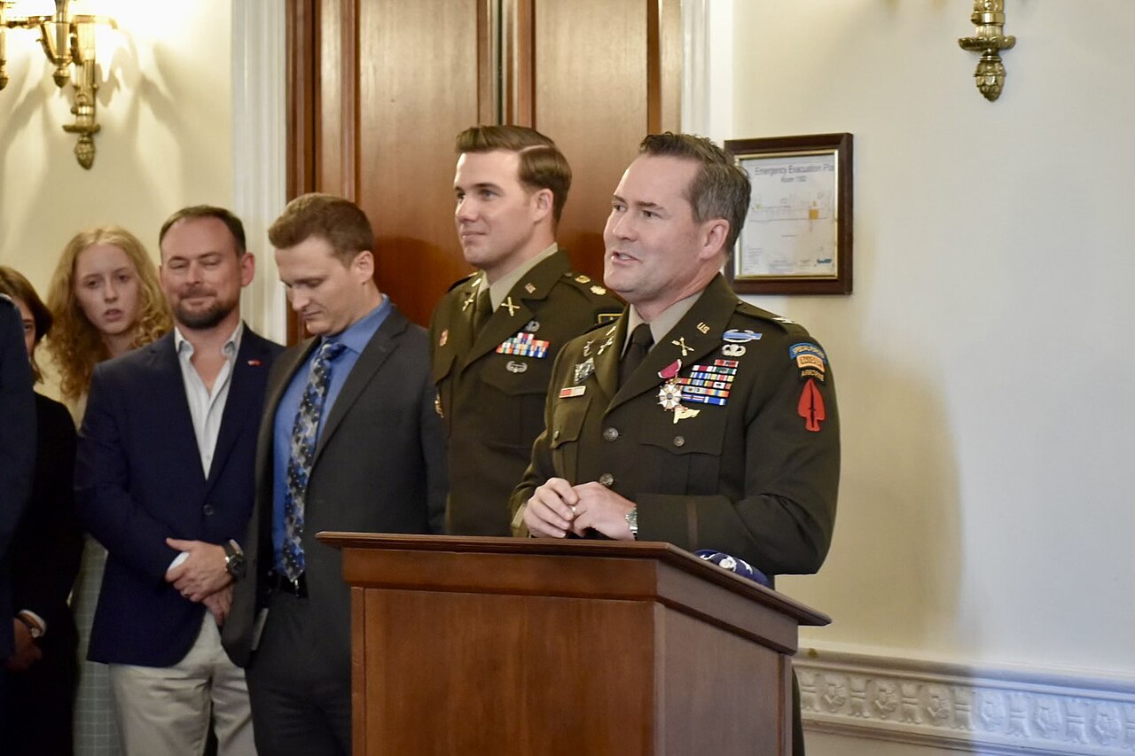 A man in a military uniform stands at a podium, speaking. Behind him, other men and a woman stand in a line.