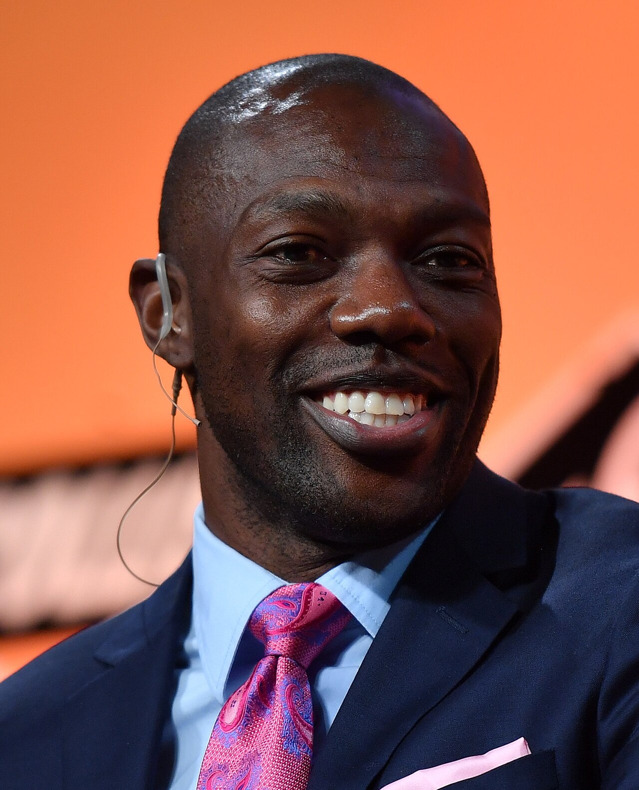 A bald black man in a blue suit, pink patterned tie, and blue shirt smiles, showing teeth, with a white wired earpiece in his left ear.