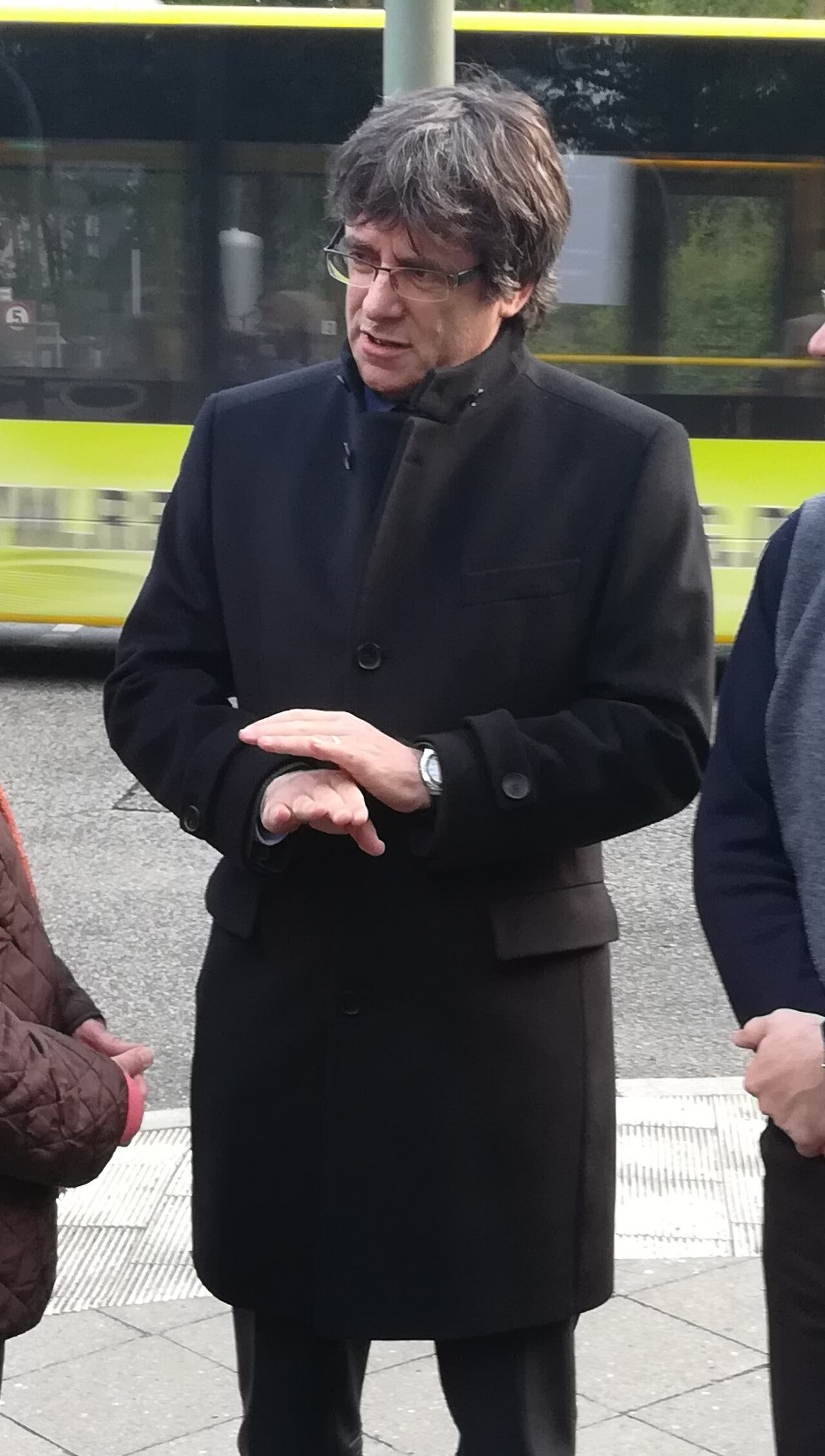A man with short, graying hair and glasses wearing a long black coat stands with his hands clasped in front of him.