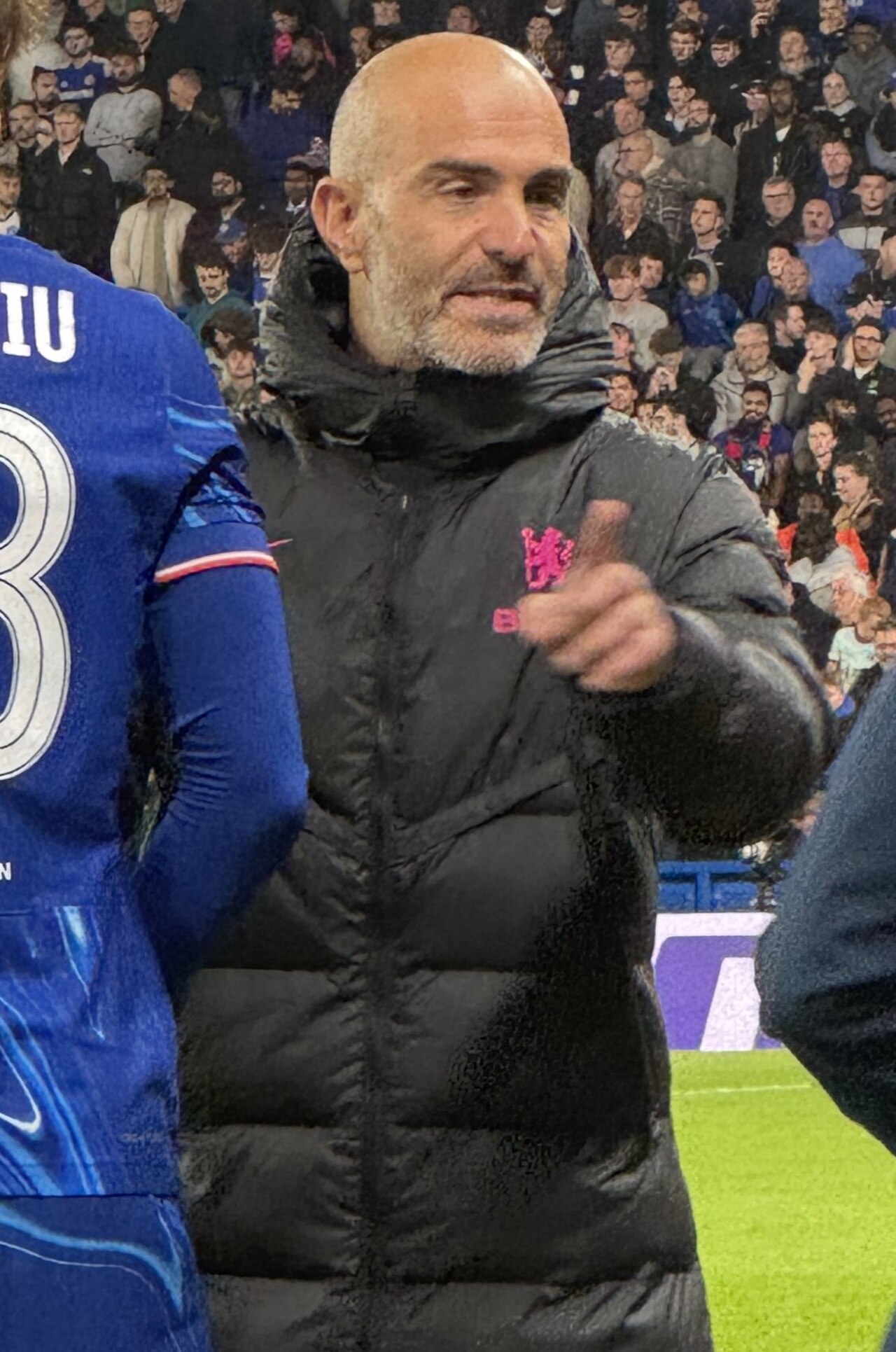 A bald man with a gray beard wearing a black puffer jacket with a pink logo on the left side of his chest stands on a soccer field, gesturing with his right hand.