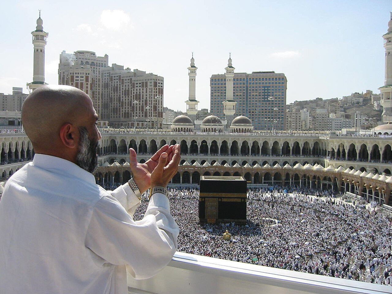 A man with a beard and bald head prays with his hands raised in front of the Kaaba in Mecca, surrounded by a large crowd of people.