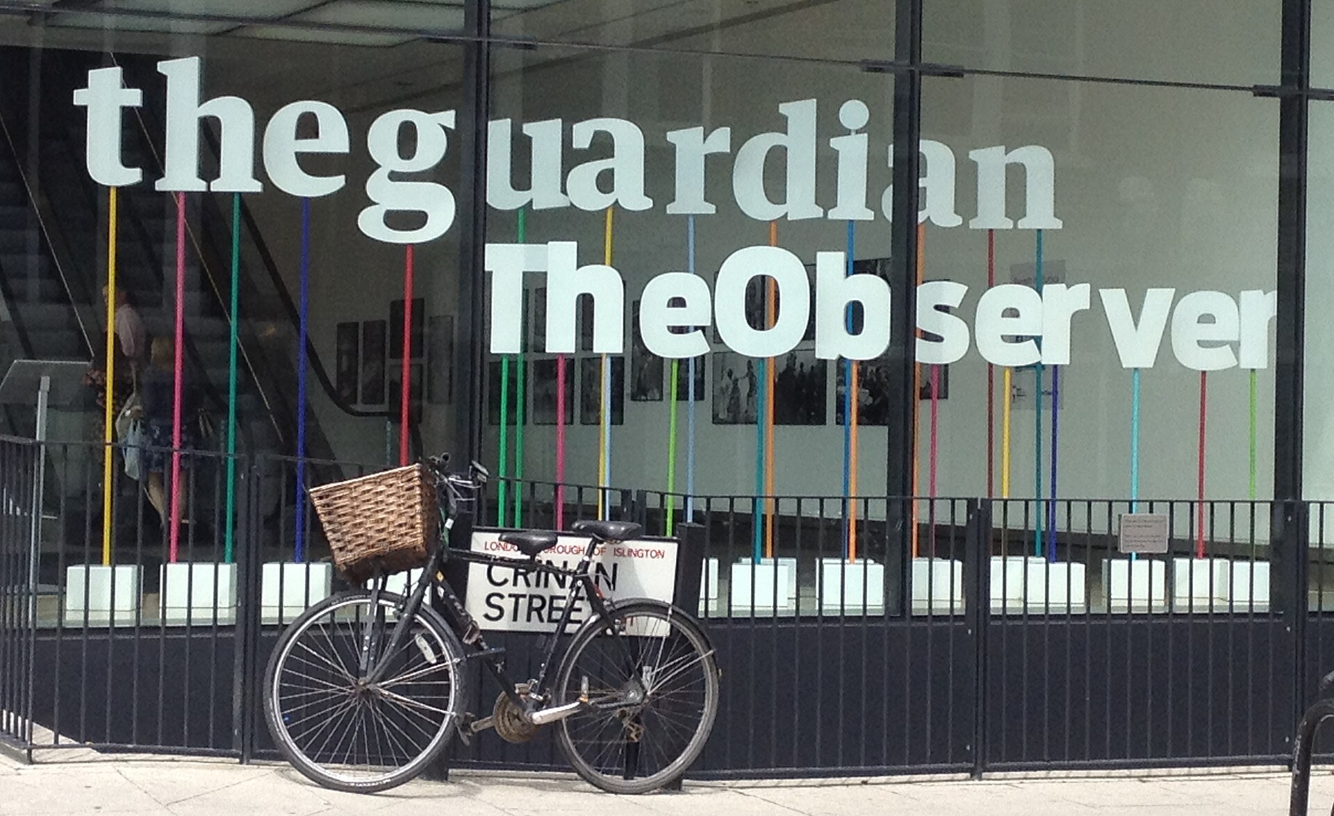 The words "the guardian TheObserver" are displayed in large white letters on a glass window. Below the text, colorful vertical lines are visible, and a bicycle with a basket is parked in front of the window.
