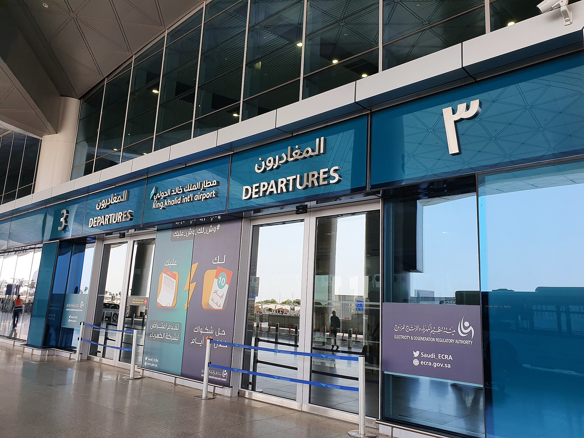 The exterior of King Khalid International Airport's departures terminal, with Arabic and English signage above glass doors.