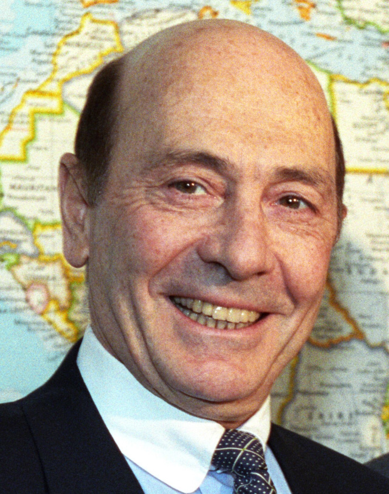 A smiling older man with a balding head, wearing a dark suit, white shirt, and patterned tie, in front of a map background.