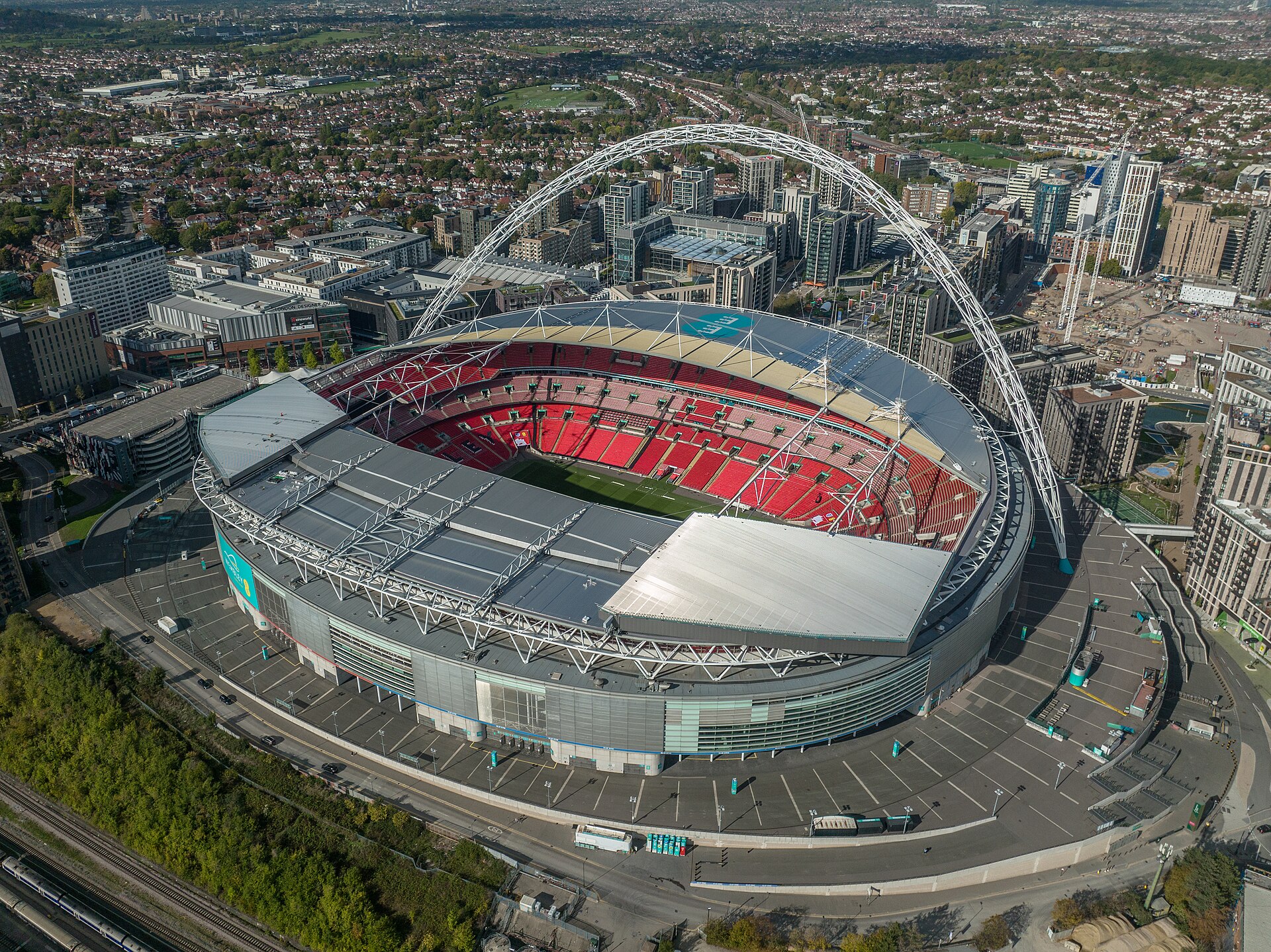 An aerial view of Wembley Stadium, a large football stadium with a distinctive arch, surrounded by urban development and greenery.
