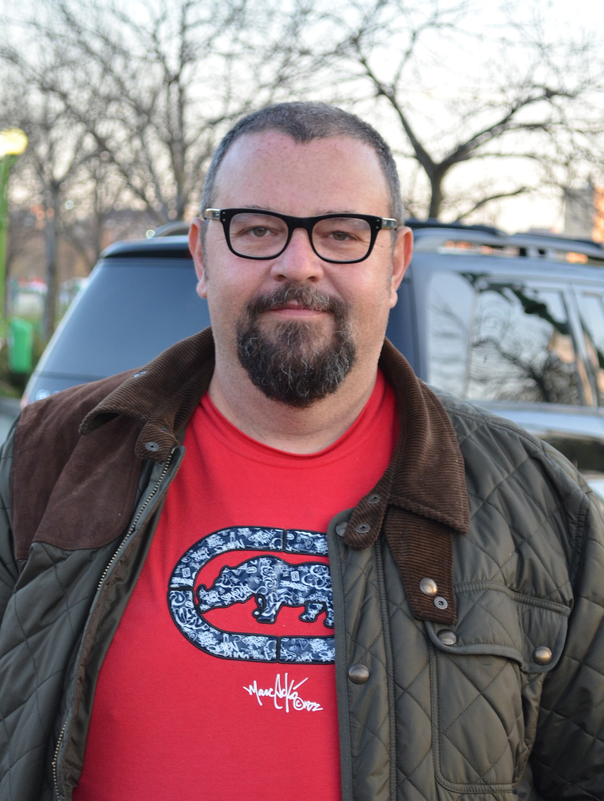 A man with short hair, a beard, and glasses wearing a red shirt and green jacket.