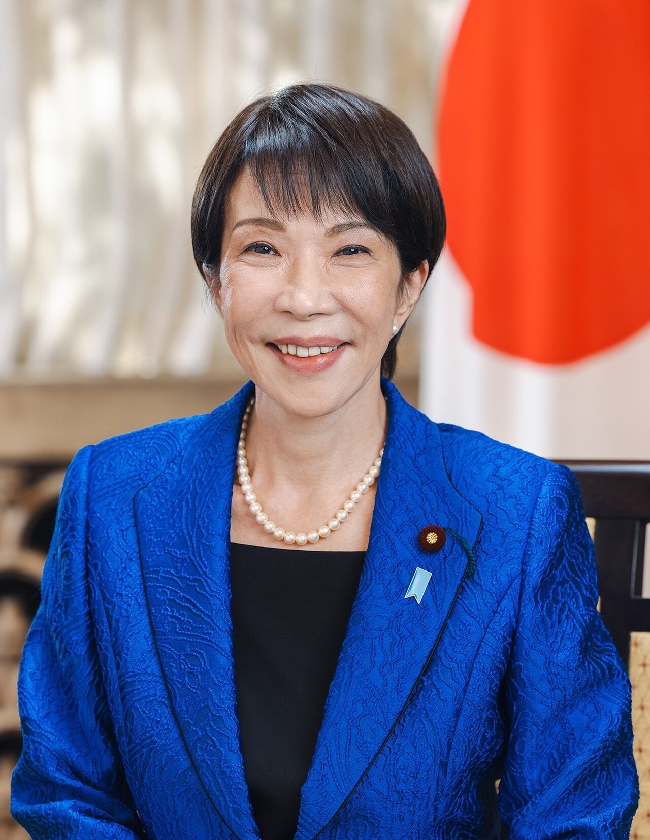 A woman in a blue blazer and pearl necklace smiles at the camera, with the Japanese flag visible behind her.