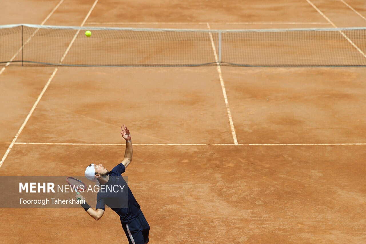 A male tennis player in a dark blue uniform and white cap serves a tennis ball on a clay court.