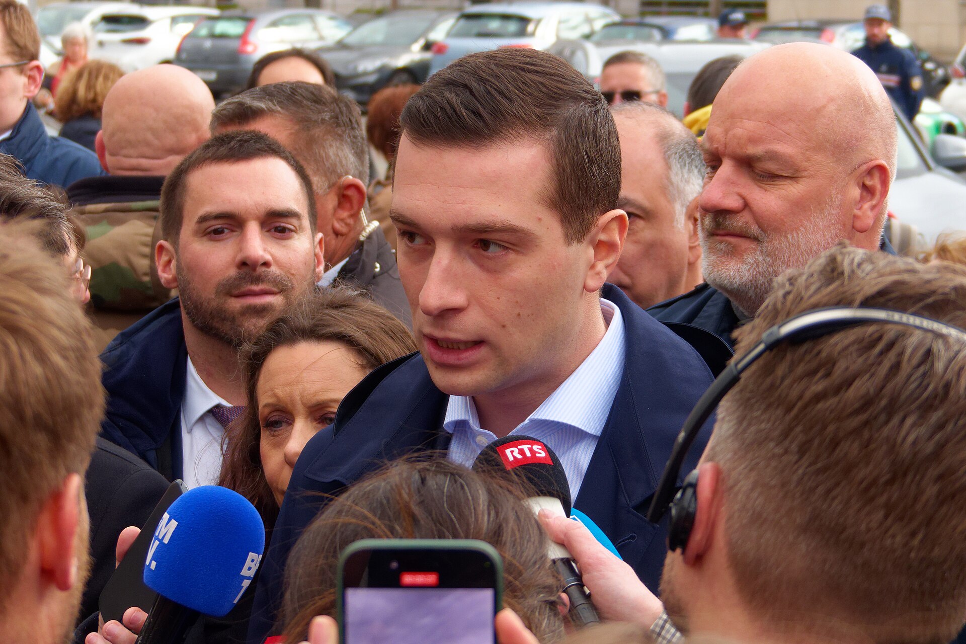 A man in a blue suit jacket and light blue shirt is speaking into microphones, surrounded by a crowd of people.