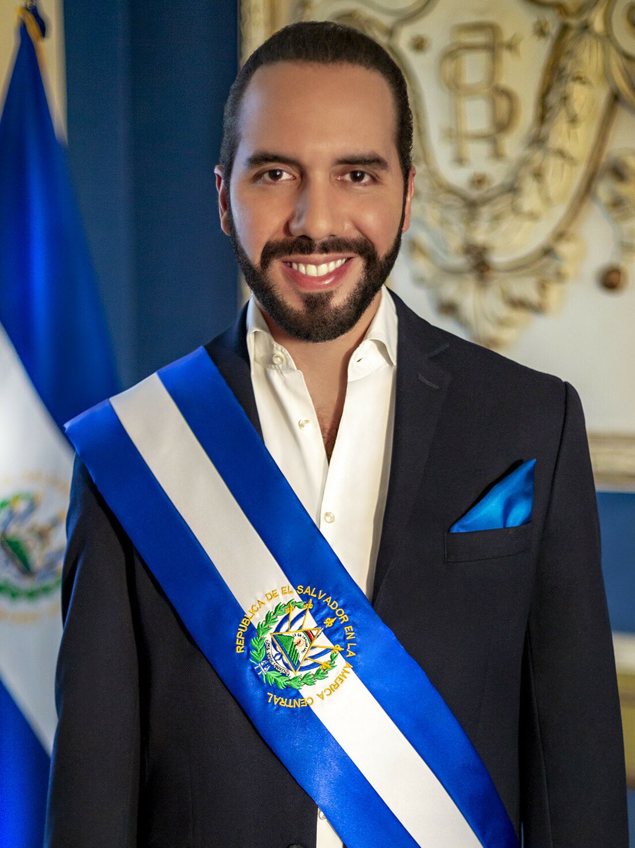 A man in a suit with a blue and white sash across his chest