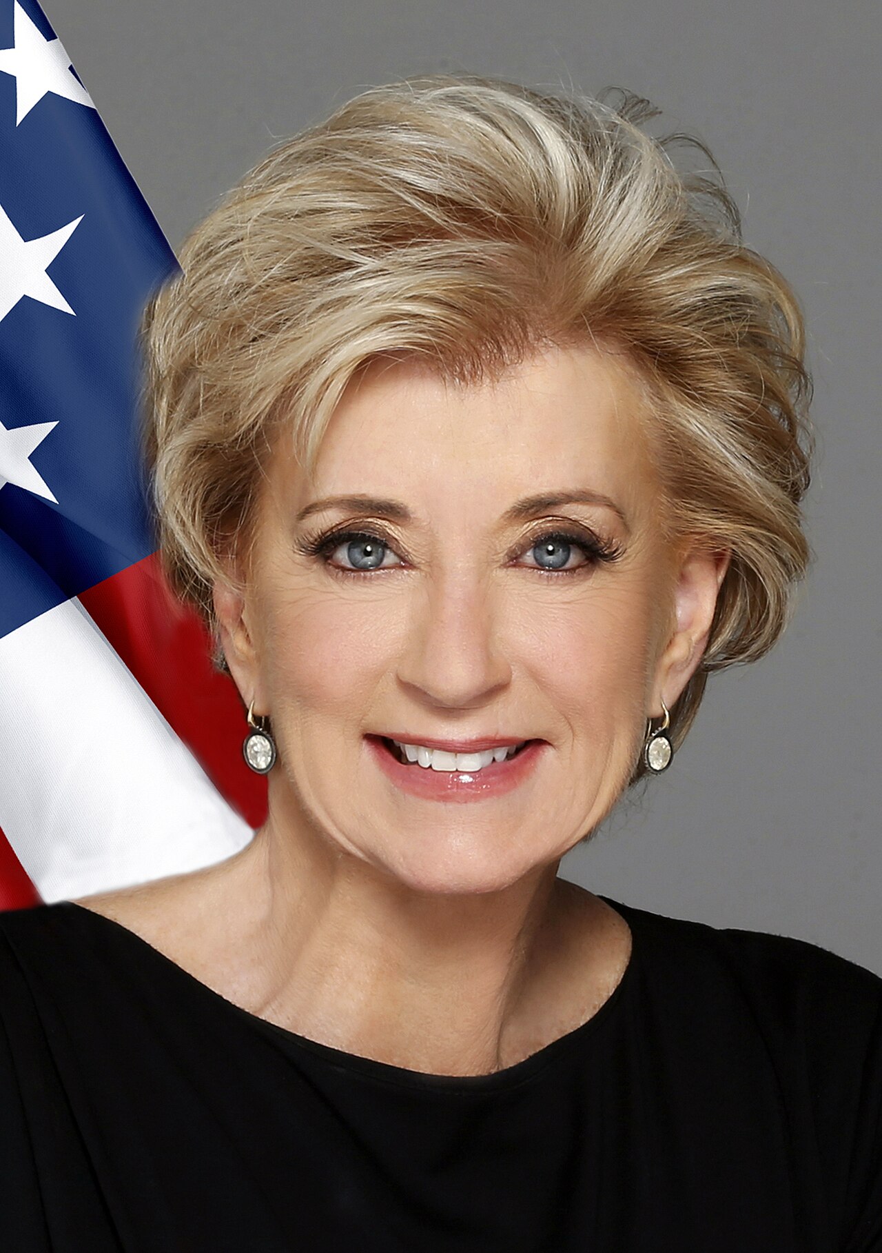 A smiling middle-aged woman with short blonde hair, blue eyes, and earrings, in front of a gray background and a partial American flag.