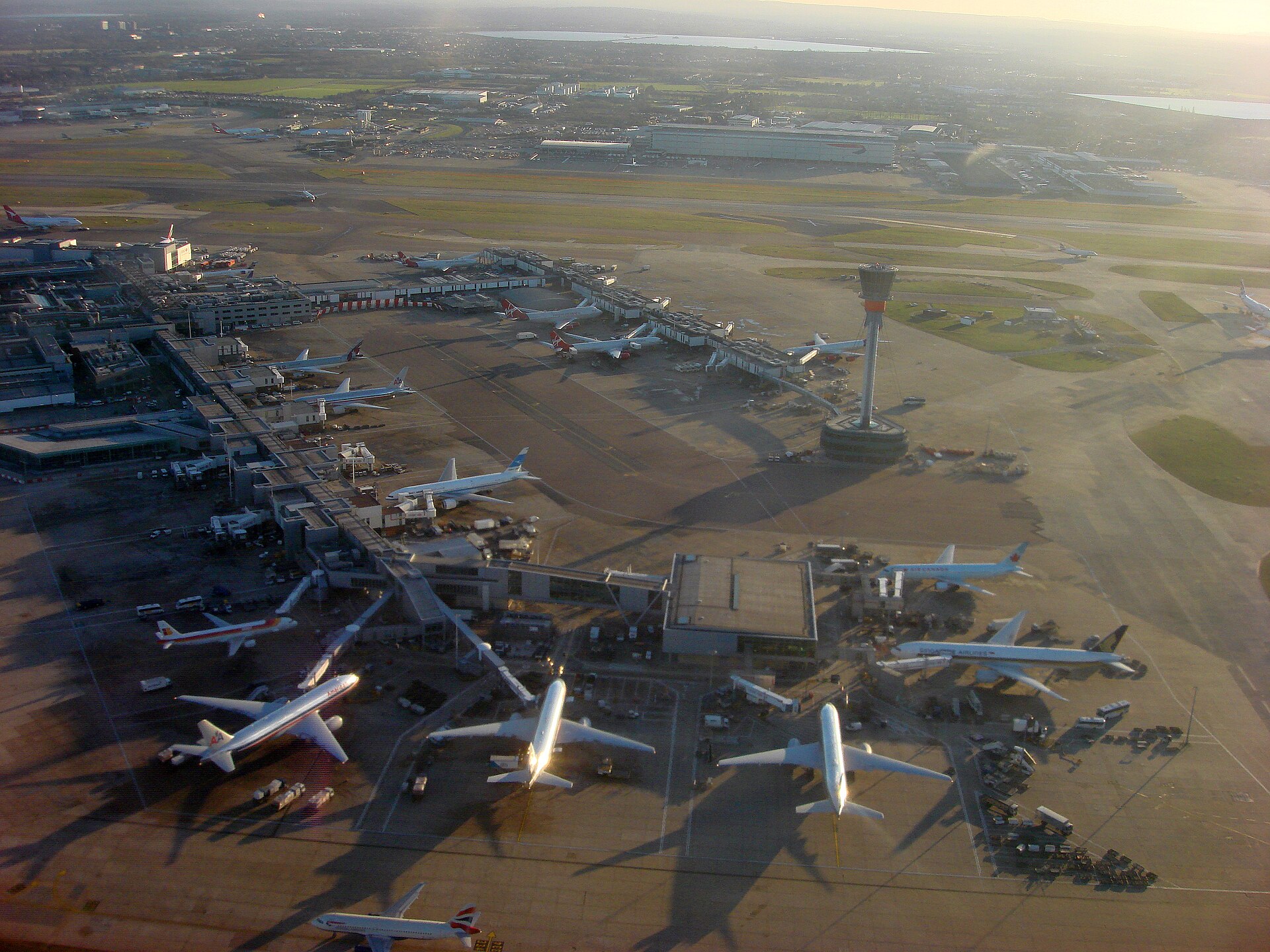 An aerial view of a busy airport with multiple airplanes parked at gates and on the tarmac. A control tower stands prominently in the middle ground.