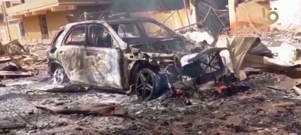 A heavily damaged and burnt-out SUV is in the foreground, surrounded by debris and rubble. The vehicle's doors are open, and the interior is scorched. In the background, a building appears to be partially destroyed.
