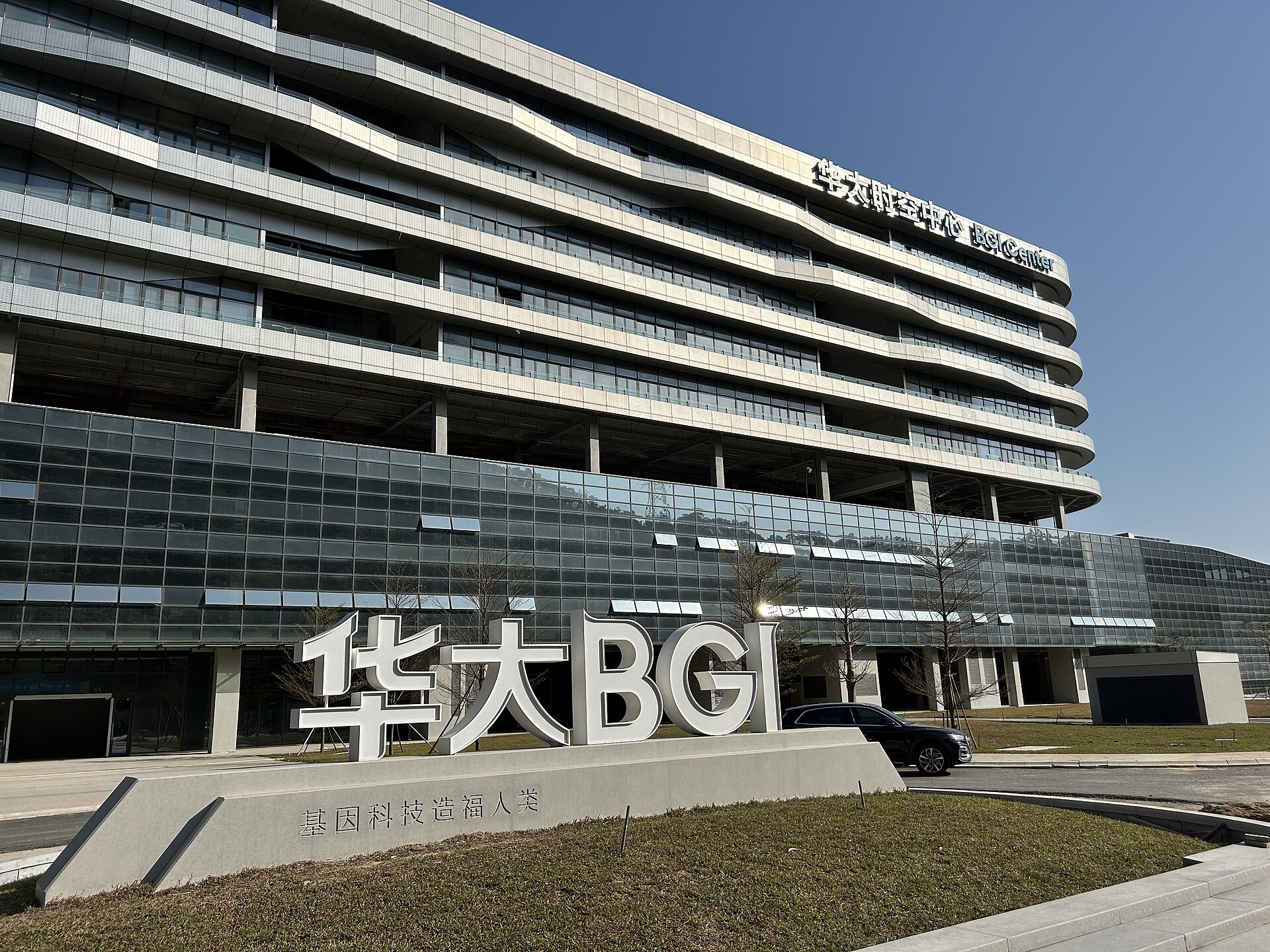 A modern, multi-story building with a glass facade and curved upper floors, featuring a large white sign in front that reads "华大 BGI" and smaller text below. A black SUV is parked to the right of the sign.