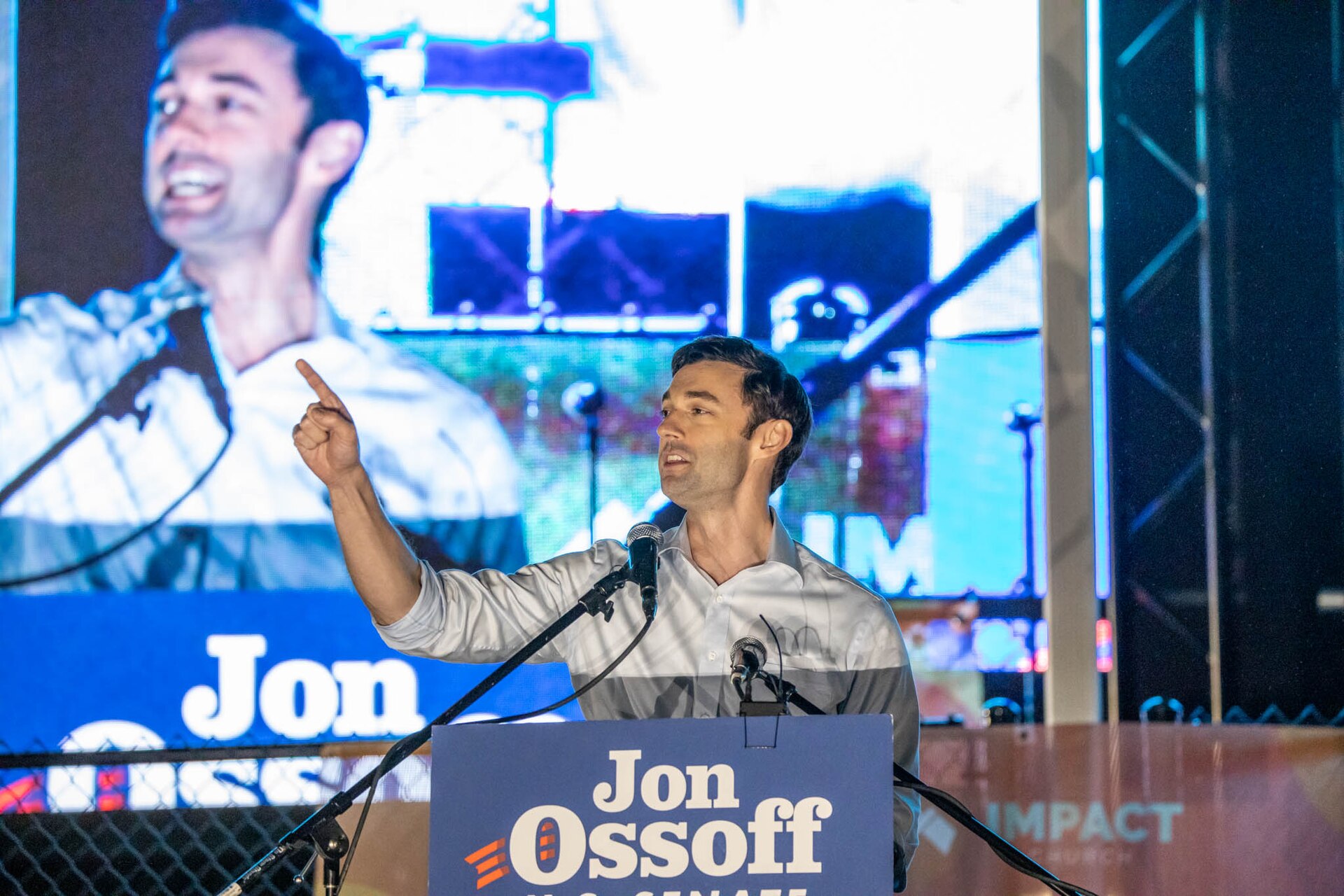 Jon Ossoff, wearing a light-colored collared shirt with gray accents, speaks at a podium with a "Jon Ossoff U.S. Senate" sign. He is pointing his right index finger upwards. A large screen behind him displays a blurred image of himself.
