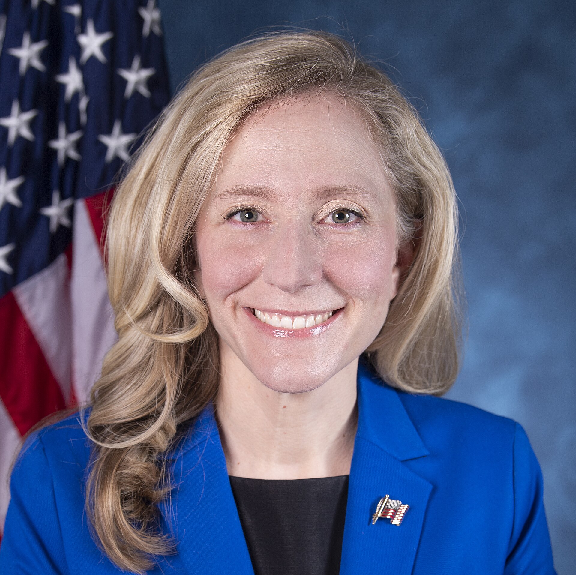 A smiling woman with blonde hair, wearing a blue blazer and a black top, stands in front of an American flag.