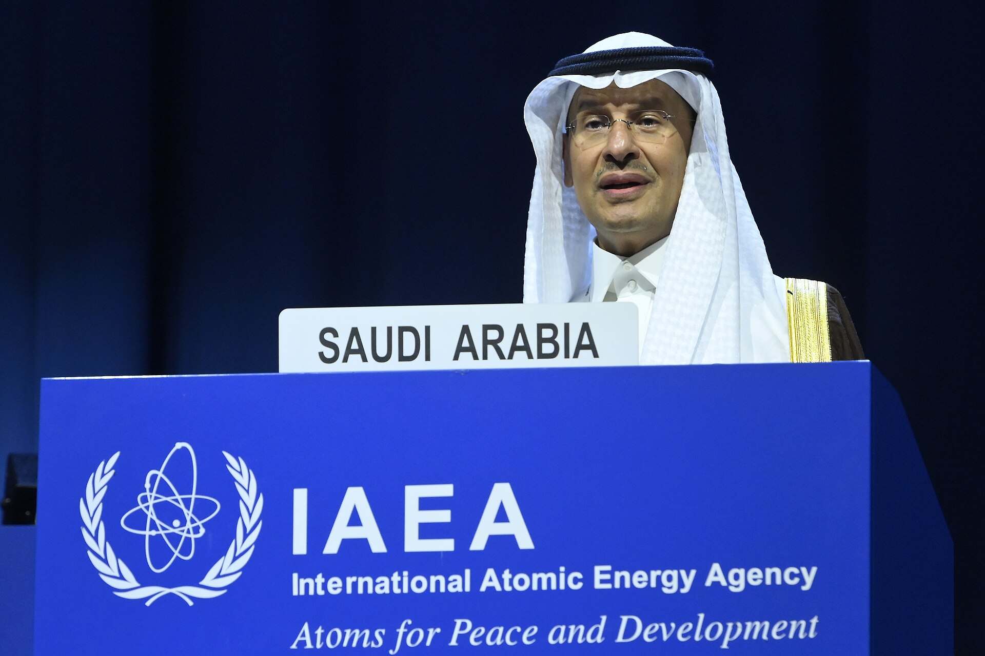 A man wearing a white robe and headscarf stands behind a blue podium with the IAEA logo and a sign that reads 'SAUDI ARABIA'.