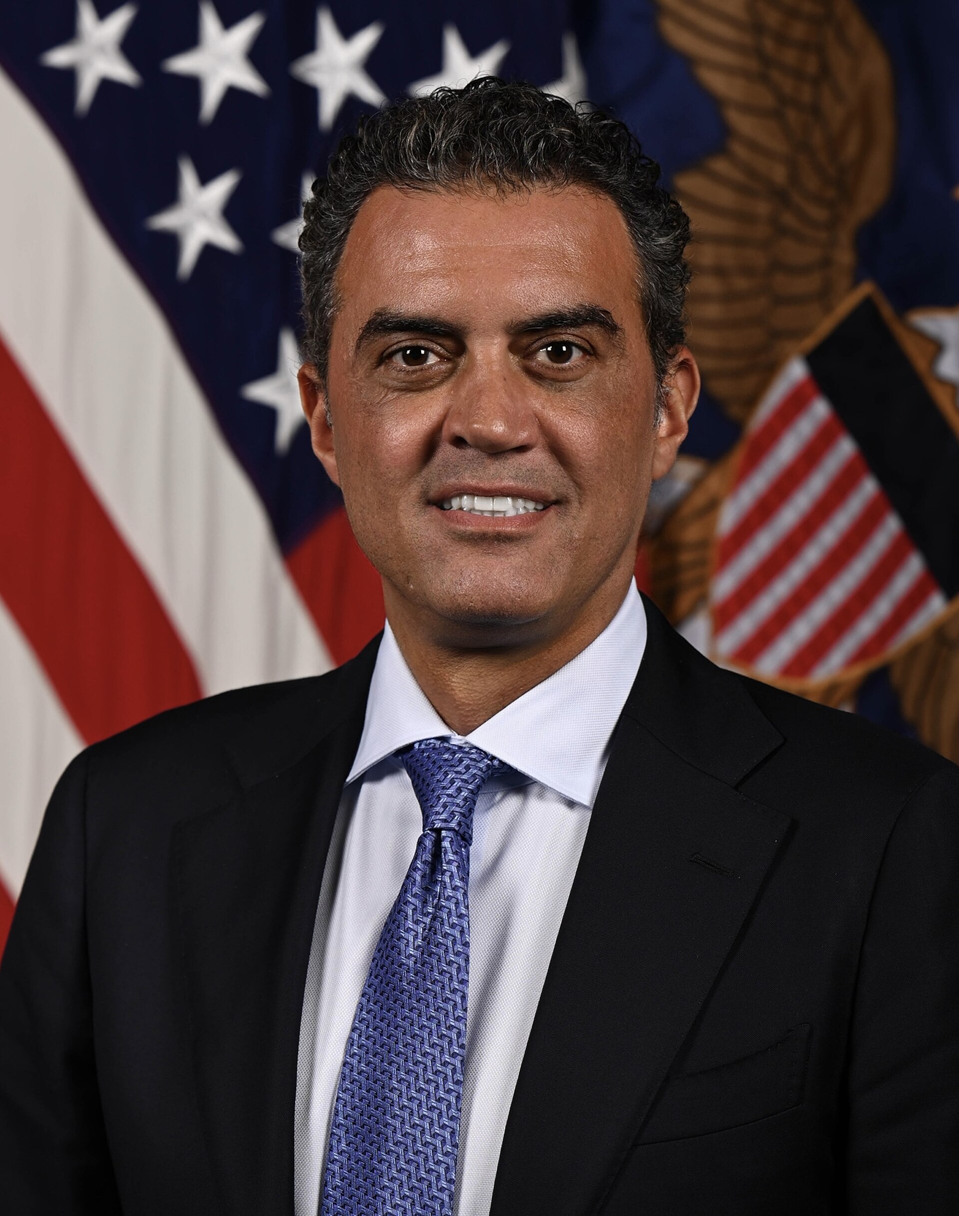 A man with short, dark, curly hair, wearing a black suit, light blue shirt, and blue tie, stands in front of an American flag and a seal.