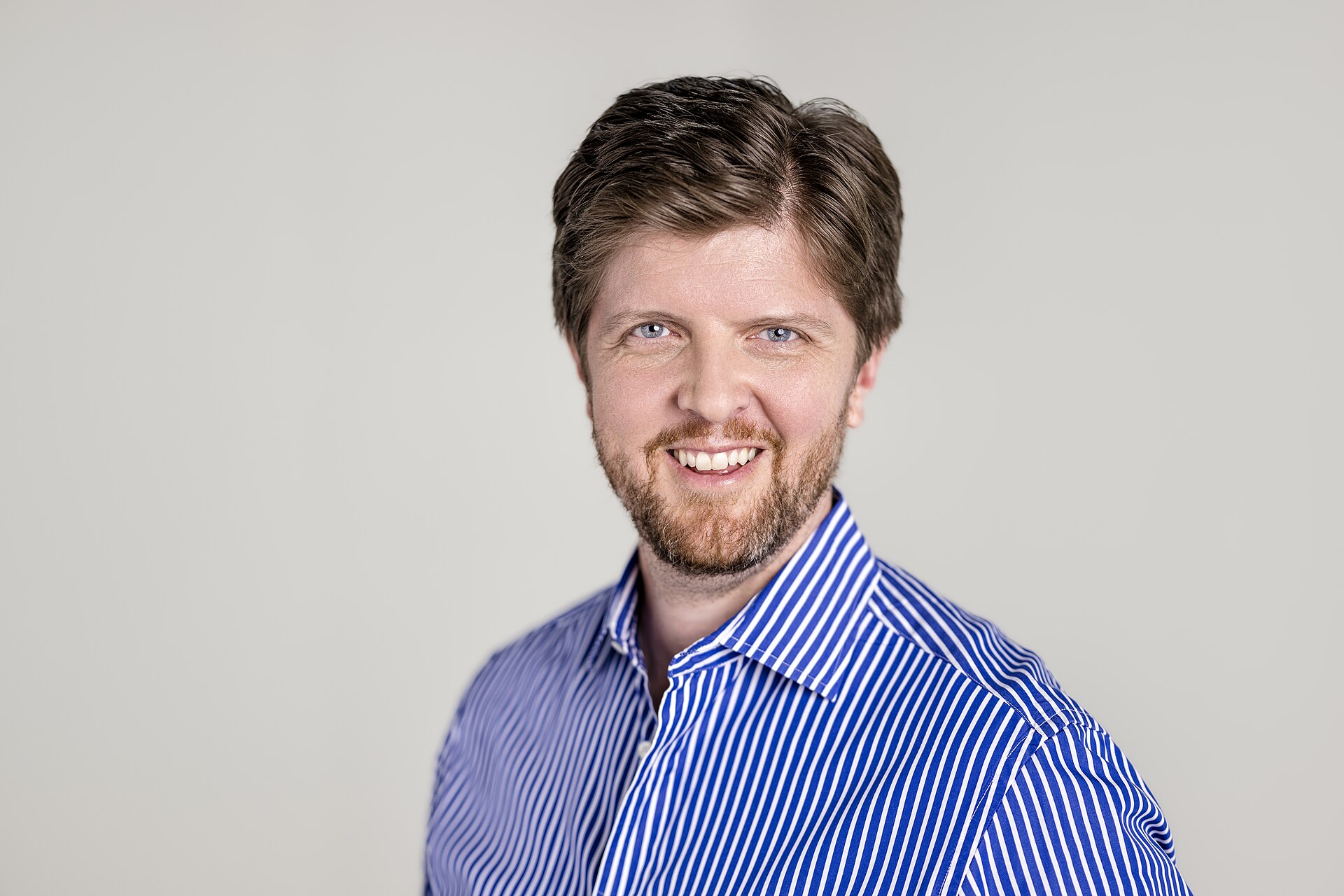 A man with brown hair and a beard wearing a blue and white striped shirt smiles at the camera.