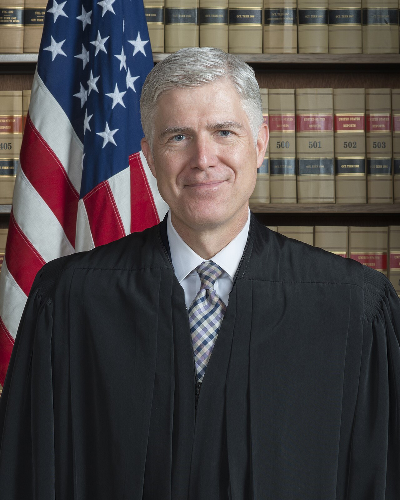 A man in a judicial robe stands in front of an American flag and bookshelves filled with law books.