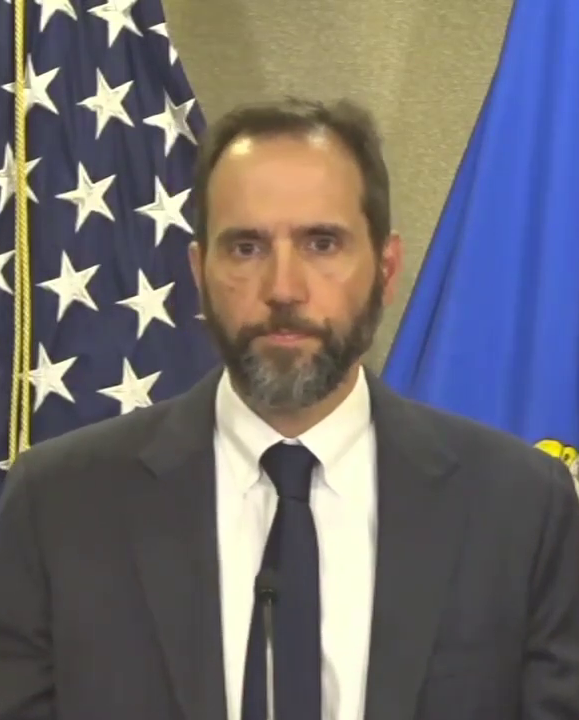 Jack Smith speaking at a Justice Department podium in 2023, with American and DOJ flags behind him.