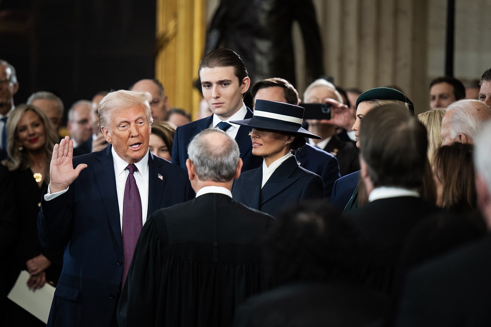Donald Trump in a navy suit, white shirt, and purple tie, with his right hand raised, standing among a crowd of formally dressed people.