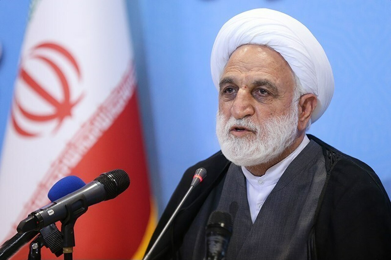 An older man with white hair and beard wearing a white turban and black robe speaking into microphones