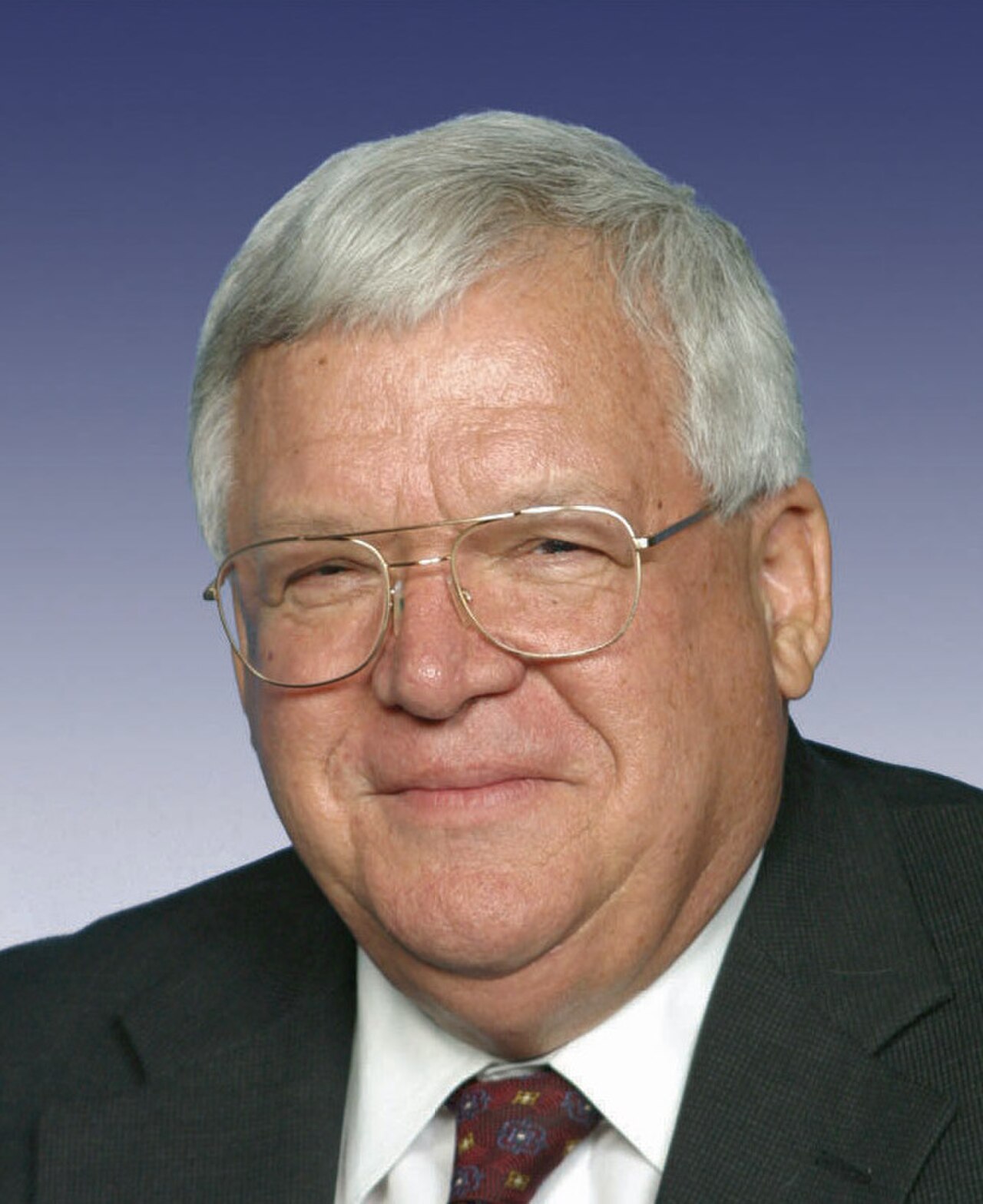 Dennis Hastert, an older white man with short gray hair, wearing glasses, a dark suit, white shirt, and patterned tie, against a blue and white gradient background.