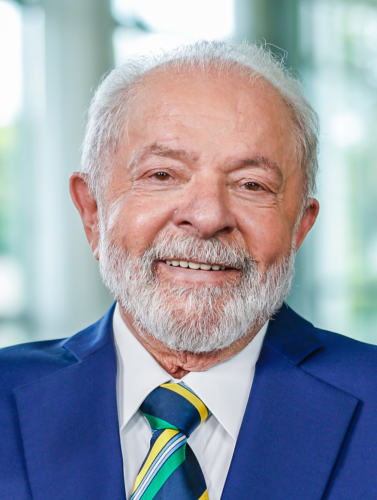 A smiling older man with short gray hair and a beard, wearing a blue suit, white shirt, and a tie with yellow, green, and blue stripes.