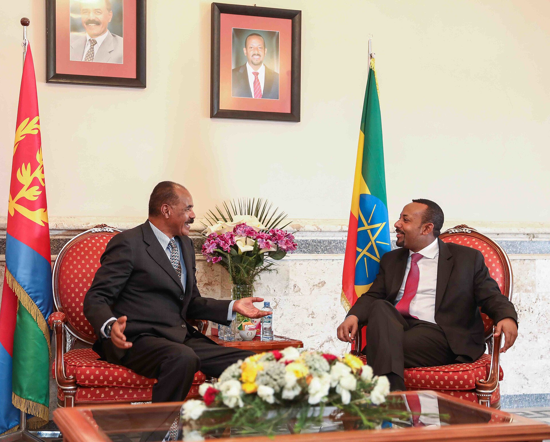 Two men in suits are seated in chairs, facing each other and talking. The man on the left is wearing a dark suit and a patterned tie, and has a mustache. The man on the right is wearing a dark suit and a red tie. Behind them are flags of Ethiopia and Eritrea, and framed portraits of leaders on the w