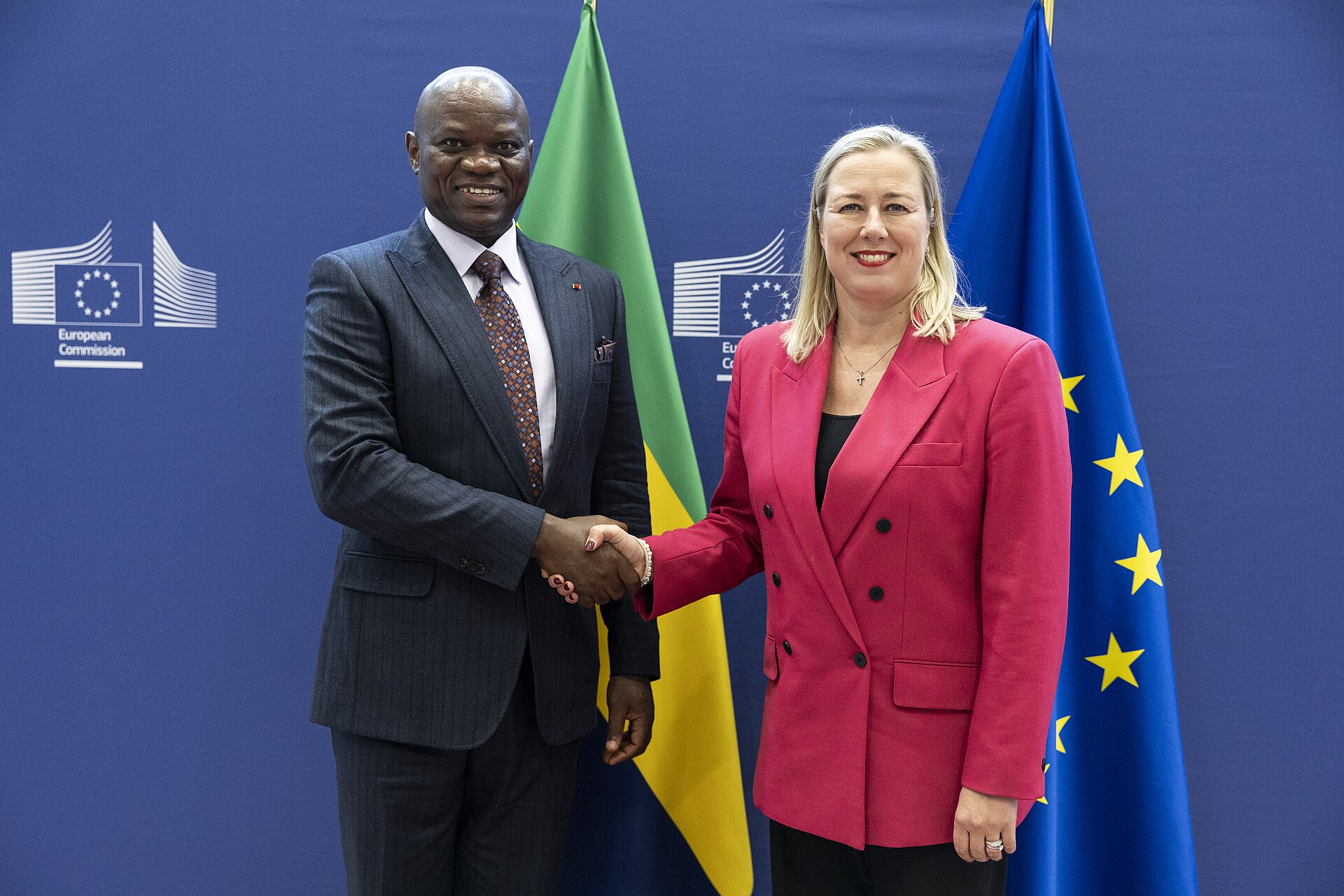 A man in a suit and a woman in a pink blazer shake hands in front of flags.