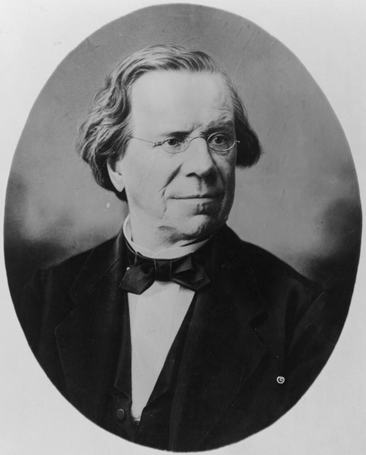 A black and white oval portrait of an older man with glasses and a bow tie.