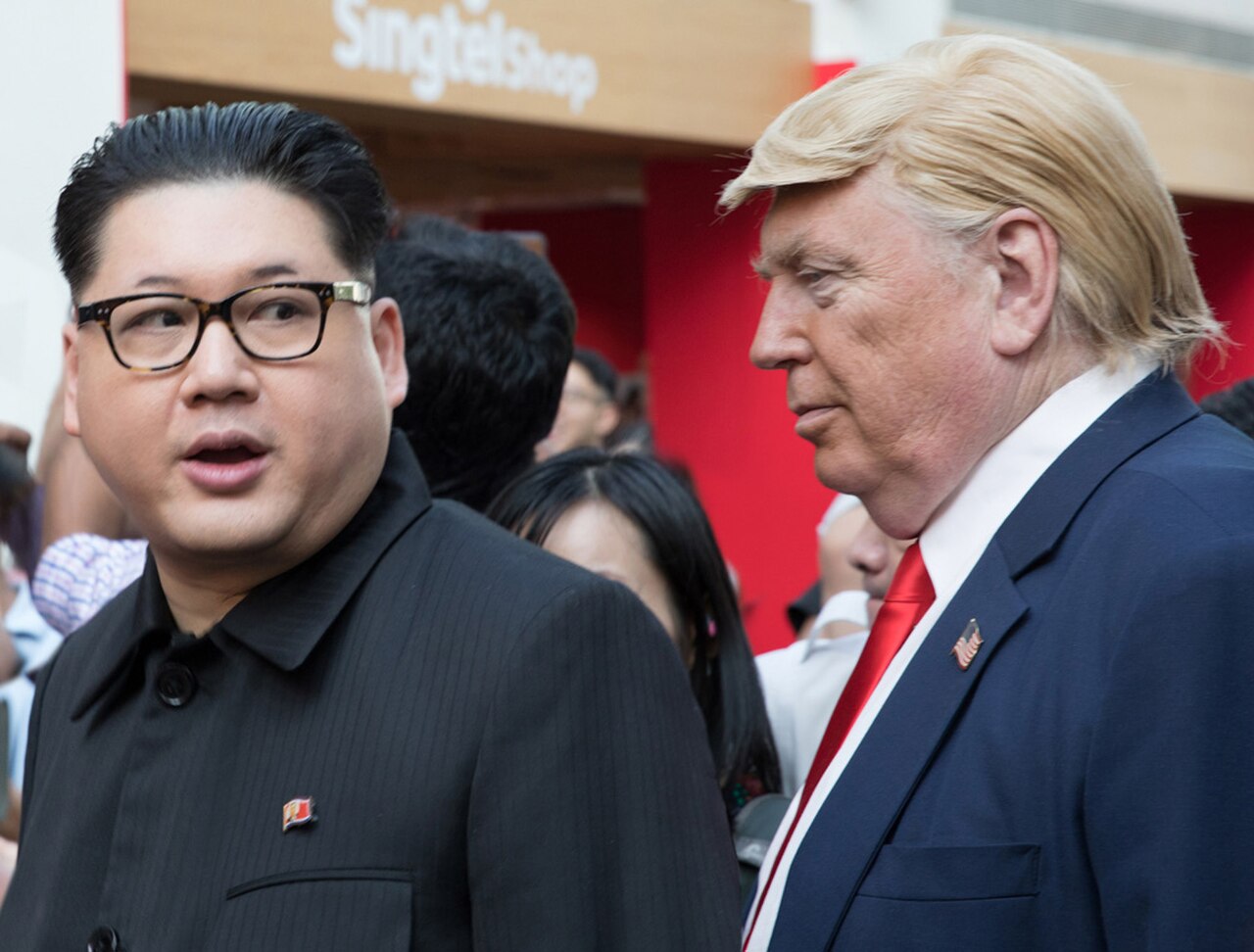 Donald Trump in a blue suit, red tie, and American flag pin, looking to the left, beside Kim Jong Un in a black suit and North Korean pin, looking forward, with a blurred crowd and Singtel shop sign in the background.
