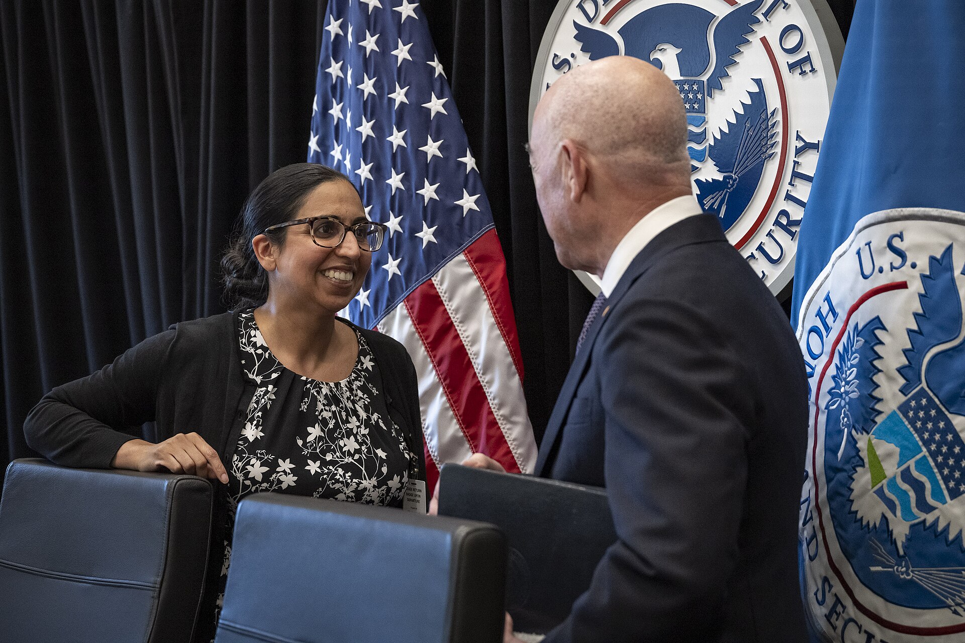 A woman with glasses and a black cardigan over a floral top smiles at a bald man in a suit, with the U.S. flag and Department of Homeland Security seals visible behind them.
