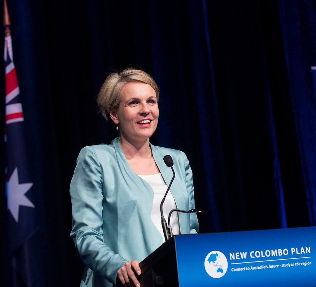 A woman with short blonde hair wearing a light blue blazer and white top stands at a podium with a blue sign that reads 'NEW COLOMBO PLAN' and 'Connect to Australia's future - study in the region'.