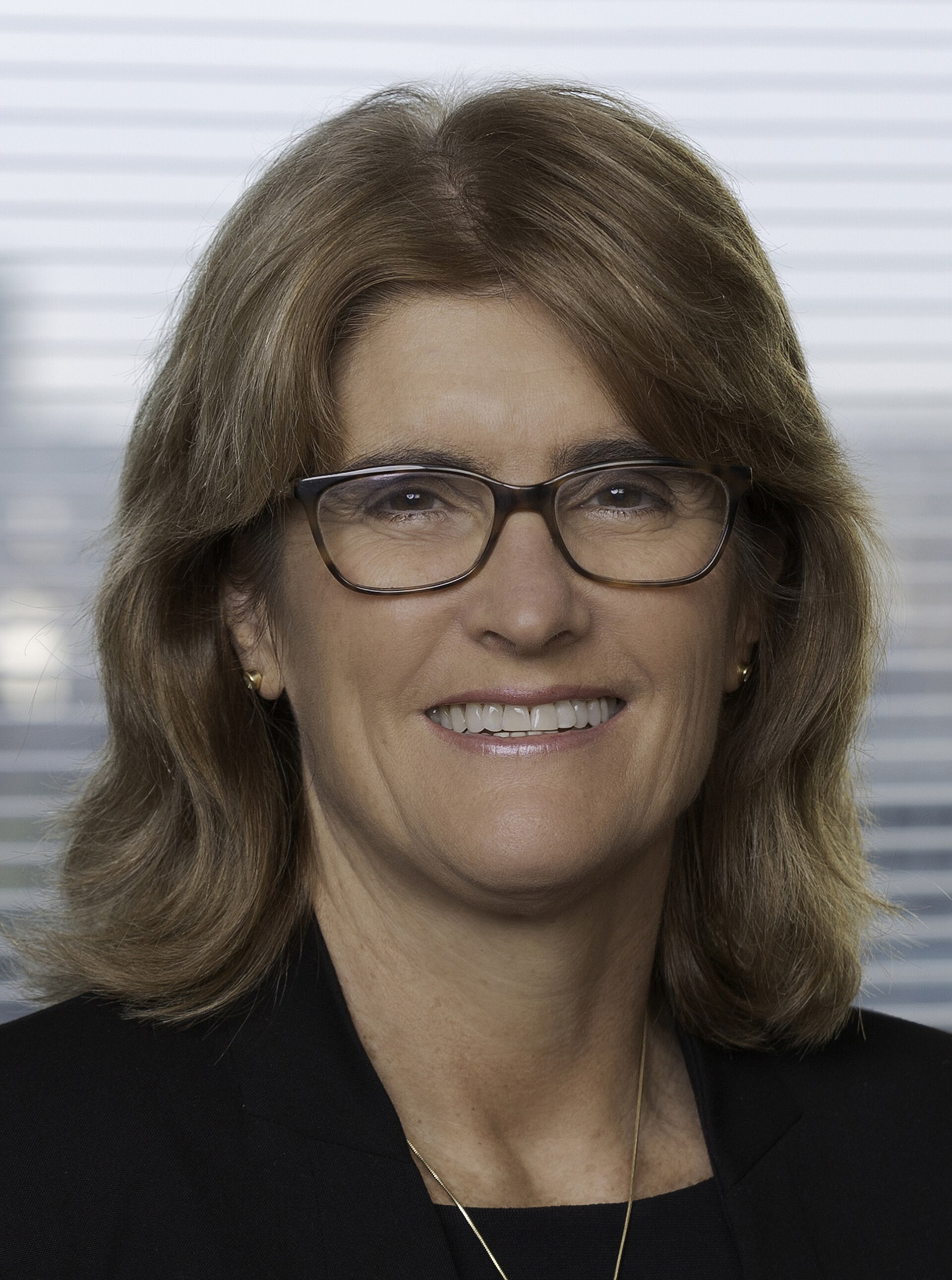 A woman with shoulder-length brown hair, wearing black-framed glasses, a black blazer, and a gold necklace, smiling at the camera.