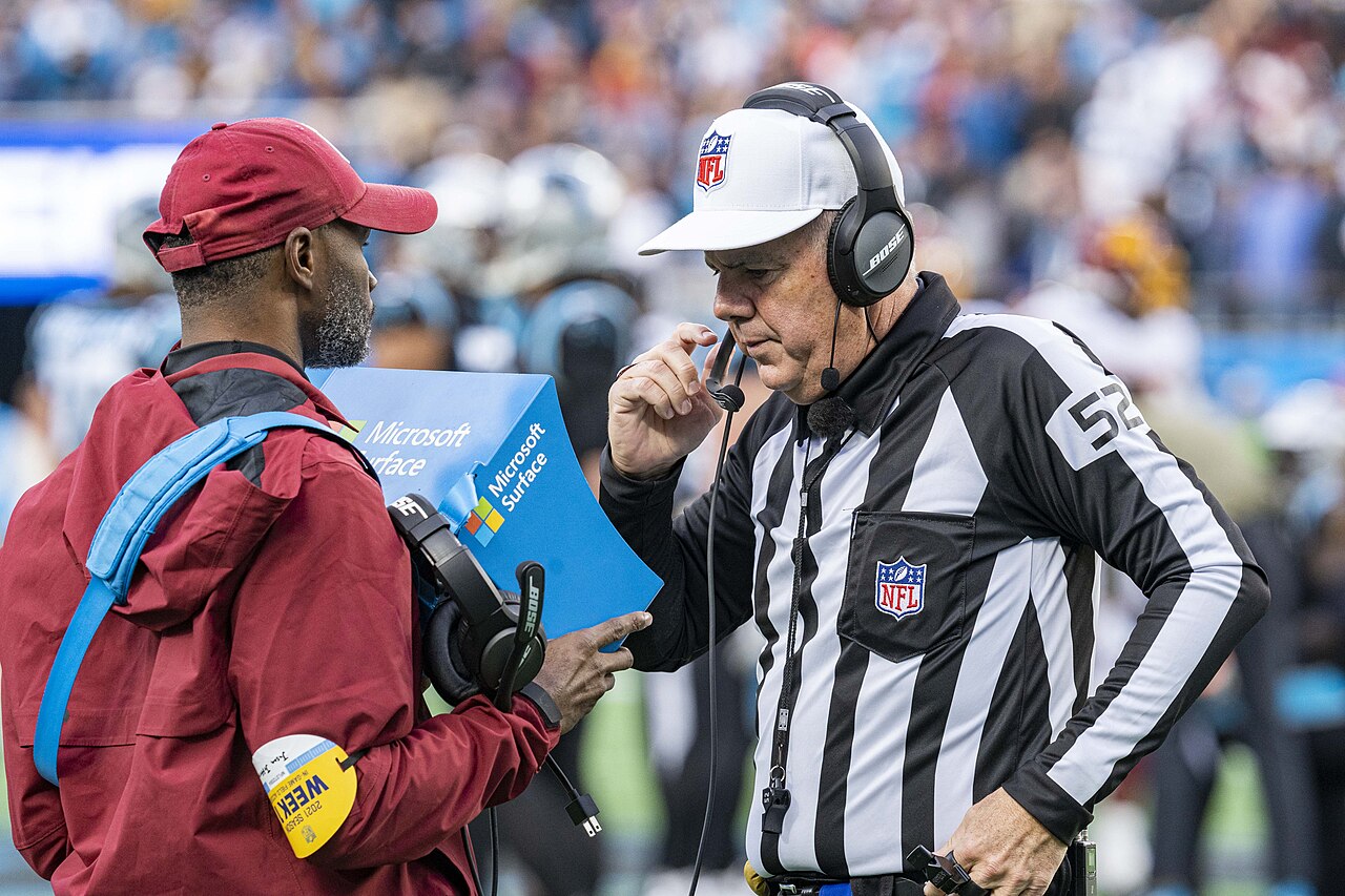 NFL referee Bill Vinovich, wearing a white cap and a striped jersey with the number 52, speaking into his headset on the sideline during a game.
