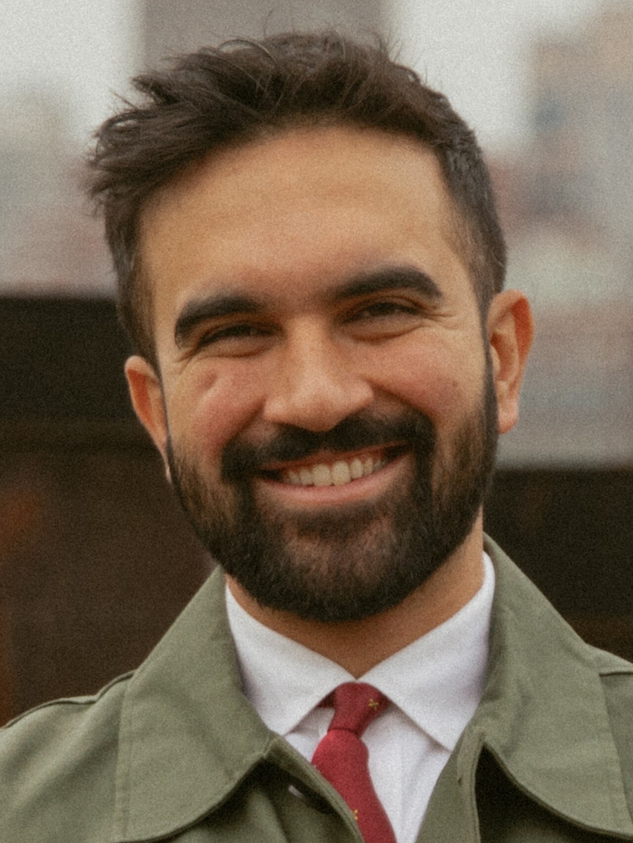 A smiling man with short dark hair and a beard, wearing a green jacket, white shirt, and red tie.