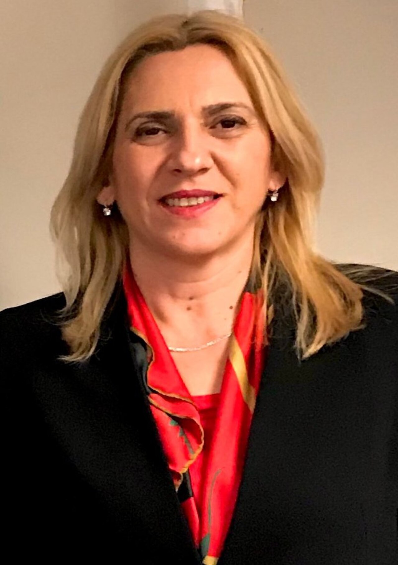 Zeljka Cvijanovic, a middle-aged woman with shoulder-length blonde hair, wearing a black blazer, red silk blouse with yellow and black accents, silver necklace, and earrings, smiling and looking directly at the camera.