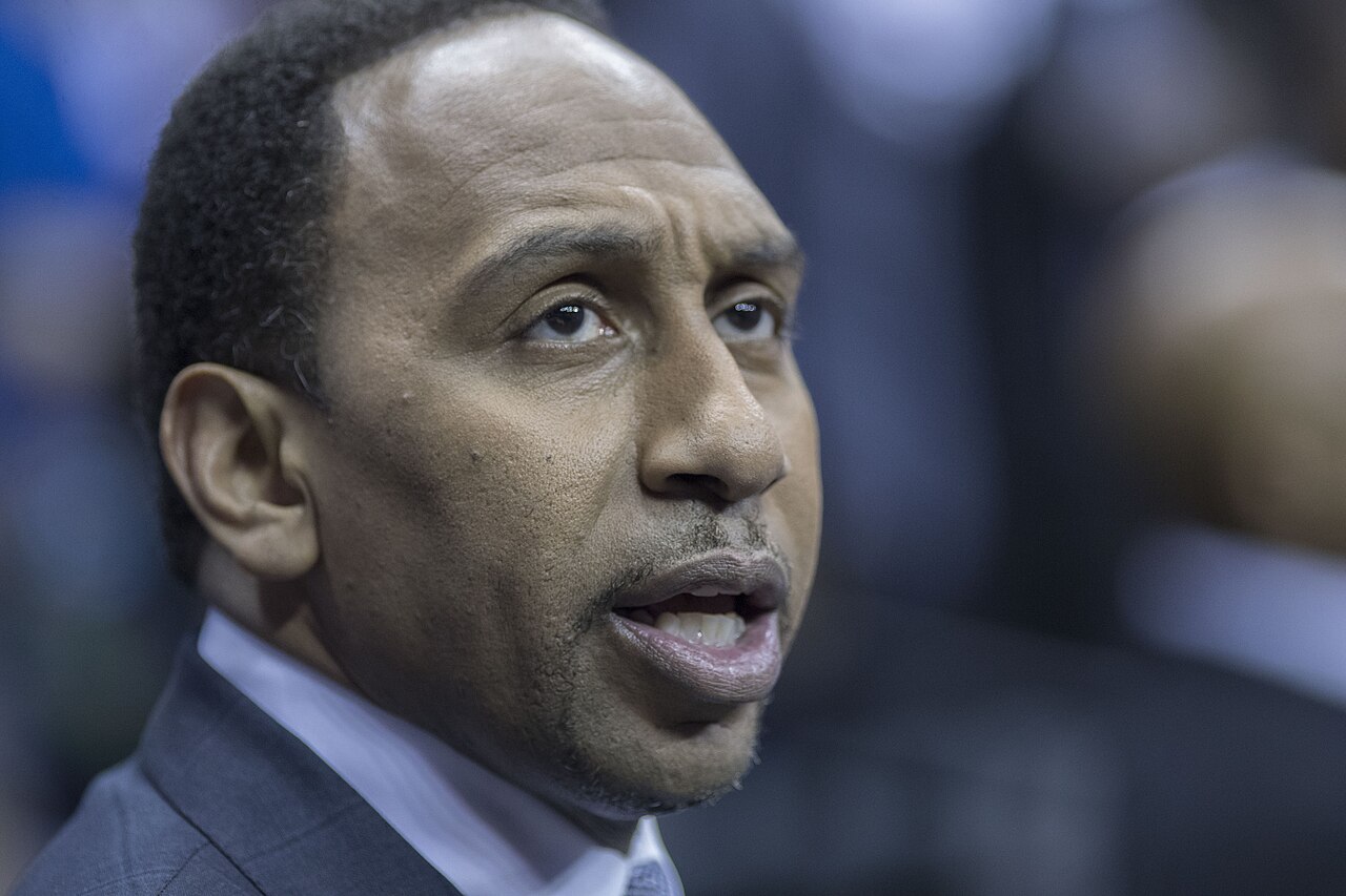 Close-up of Stephen A. Smith, a middle-aged Black man with short black hair, wearing a gray suit jacket, white collared shirt, and light-colored tie, looking to the right with his mouth slightly open.