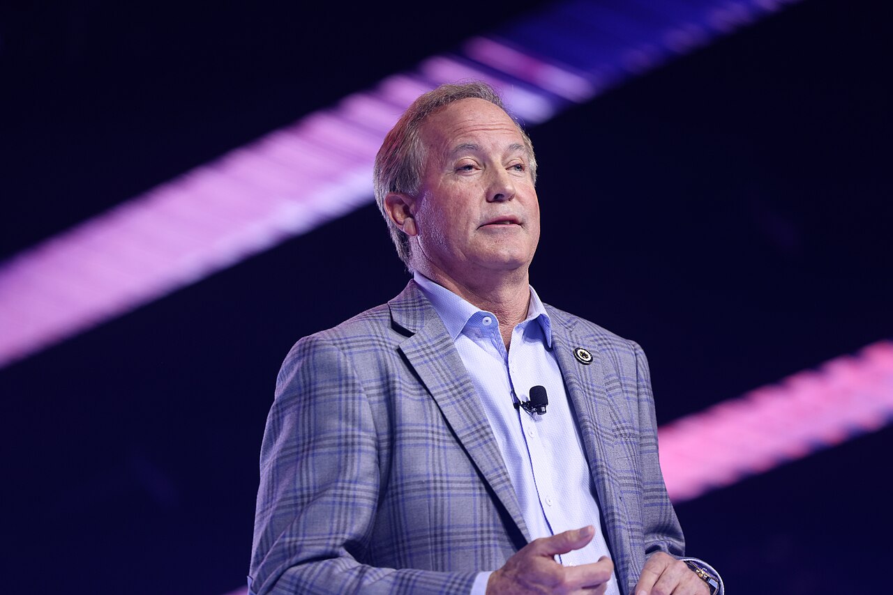 Ken Paxton, a middle-aged man with short blond hair, wearing a gray plaid suit jacket over a light blue collared shirt, with a small microphone clipped to his left lapel, gesturing with his hands in front of him.