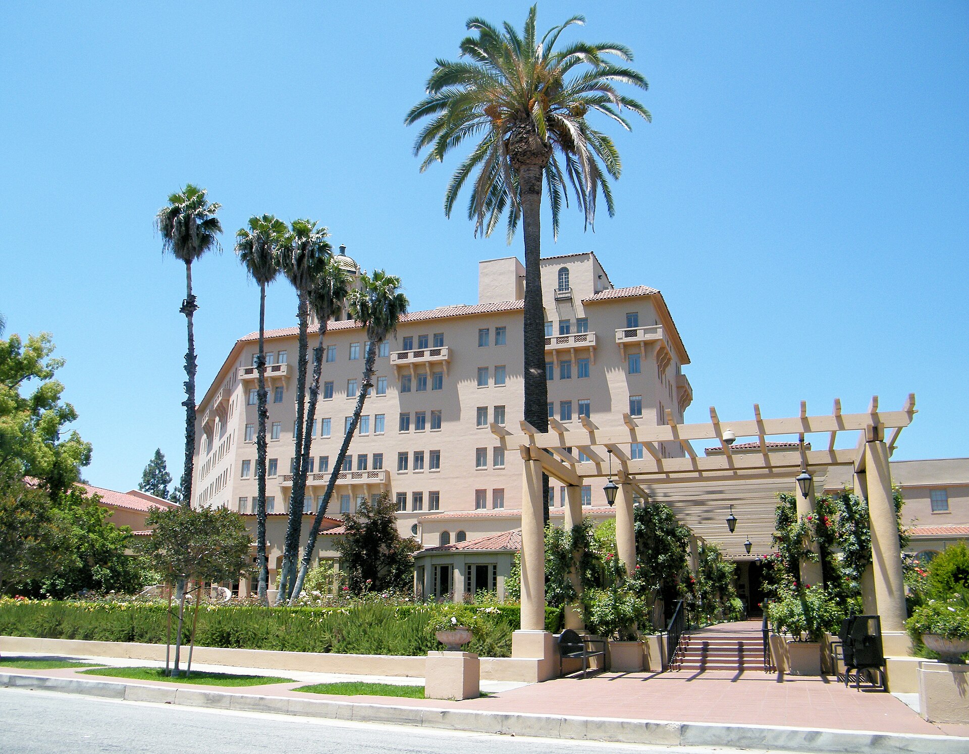 A large, light-colored building with a red tile roof and many windows is surrounded by palm trees and greenery, with a pergola and steps leading to an entrance.