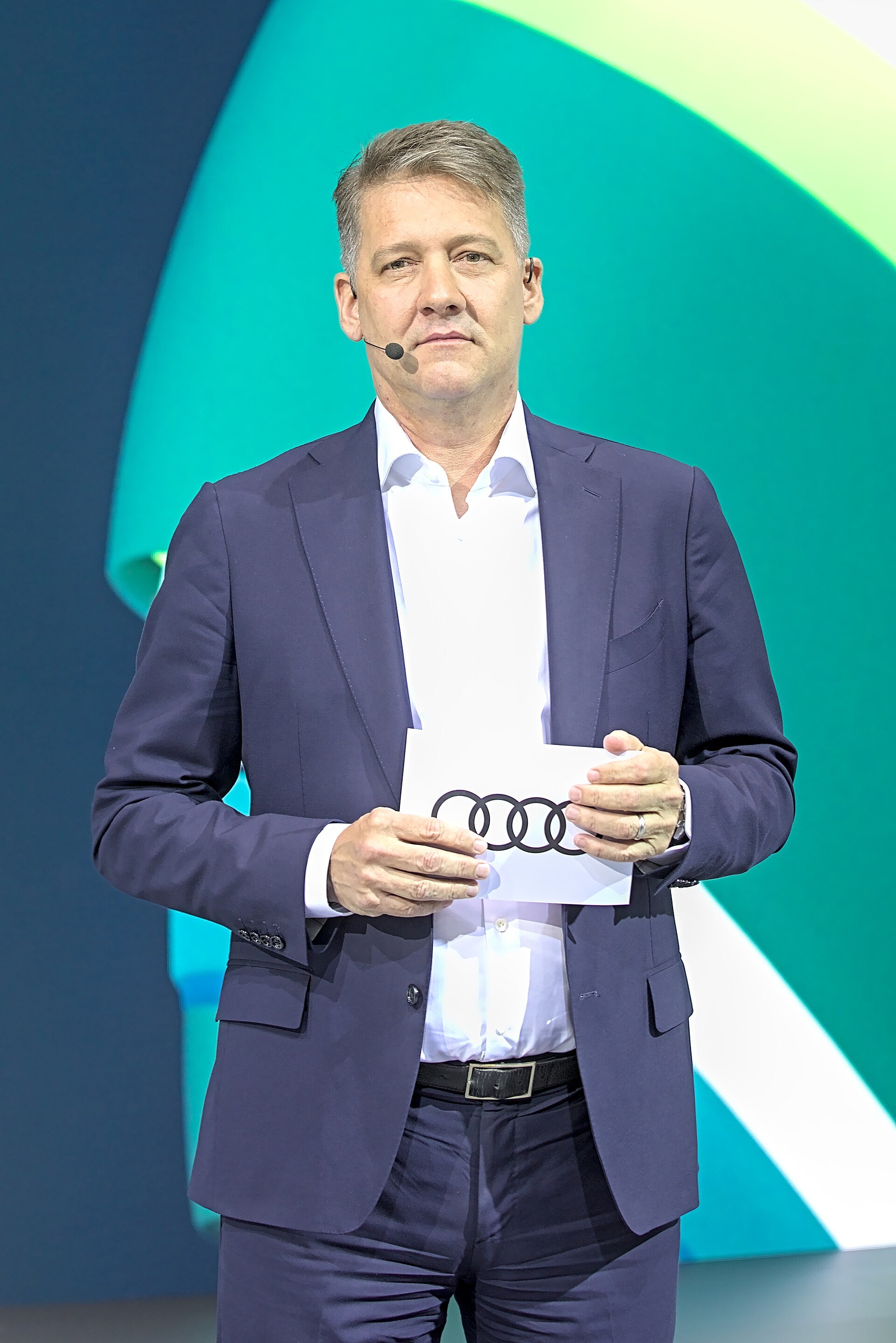 A man in a dark suit and white shirt holds a white card with the Audi logo on it.