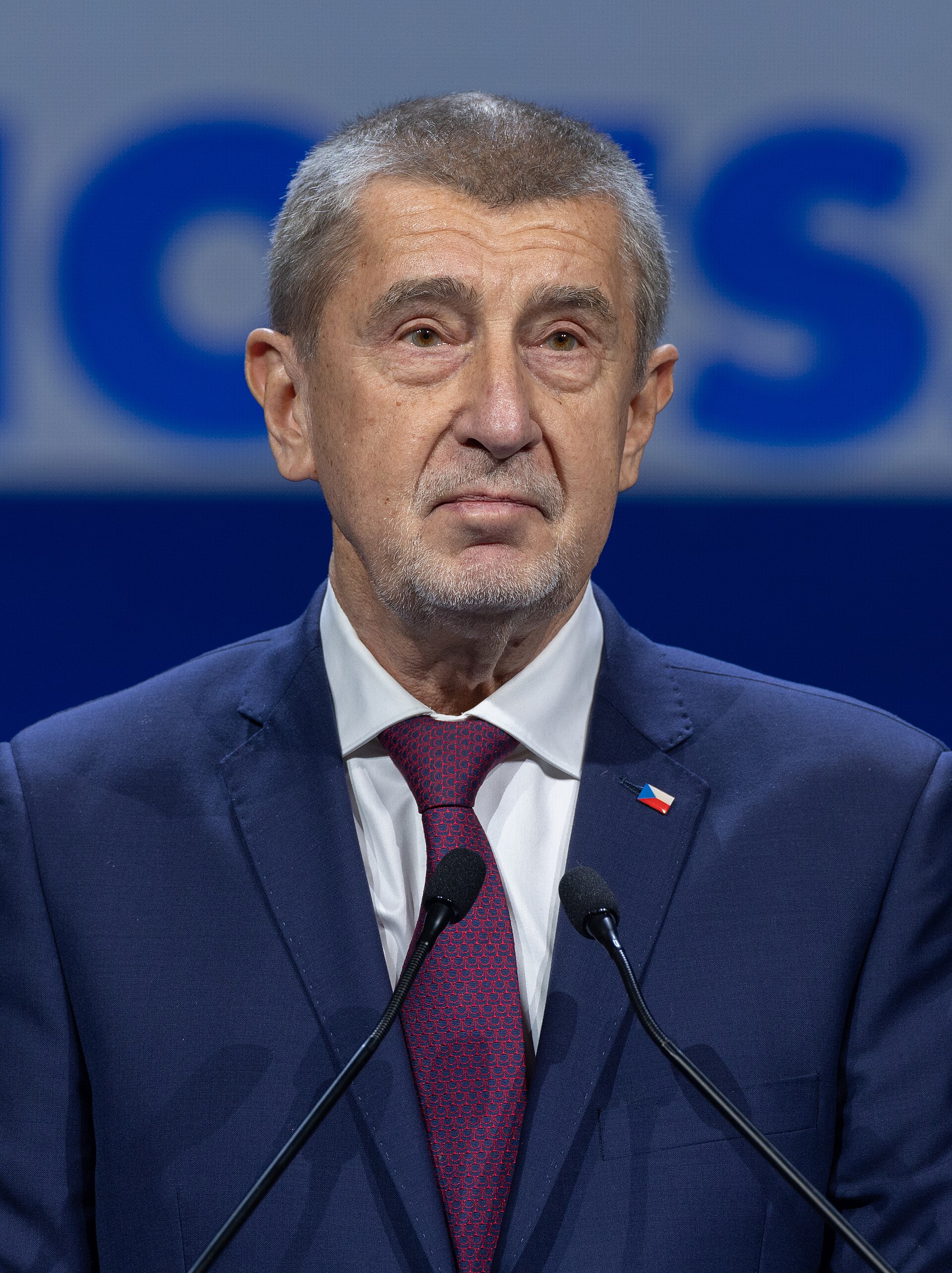 A man in a dark blue suit, white shirt, and patterned red tie stands at a podium with two microphones. He has grey hair and a goatee. A small Czech flag pin is visible on his lapel.